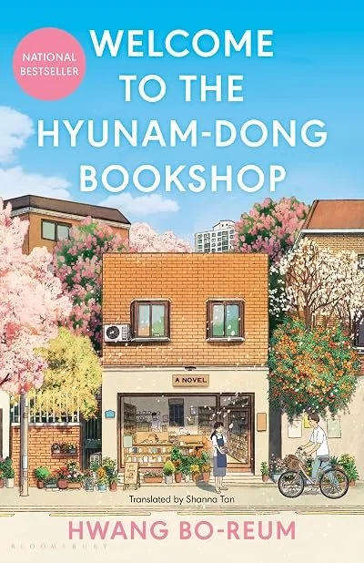 Book cover for 'Welcome to the Hyun-Dong Bookshop' by Hwang Bo-Reum, featuring a colorful illustration of a small bookstore with house-like windows, surrounded by blooming trees, with people and a bicycle outside.