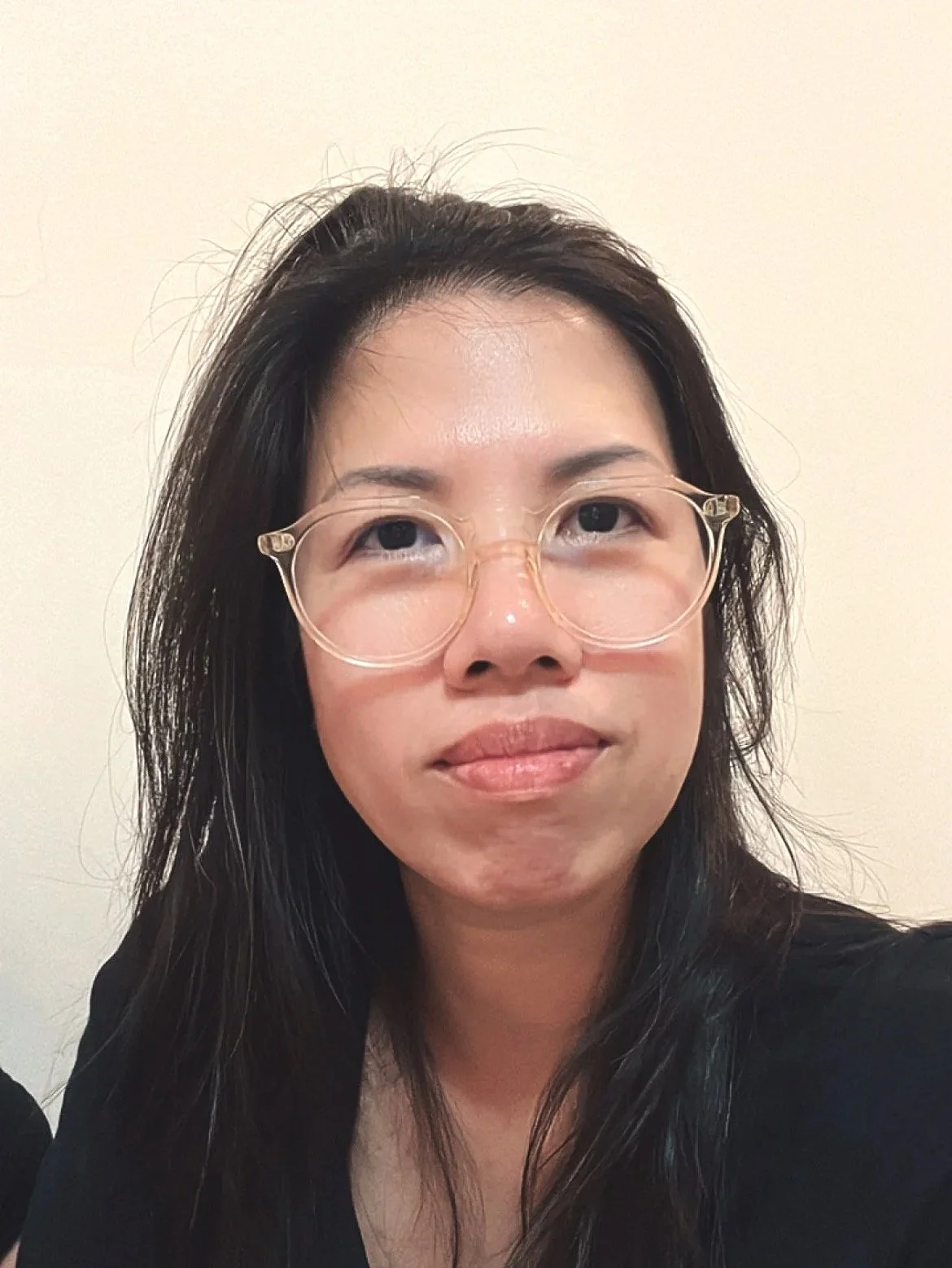 A woman with long dark hair, wearing clear round glasses, and a black top, standing against a plain light-colored wall.