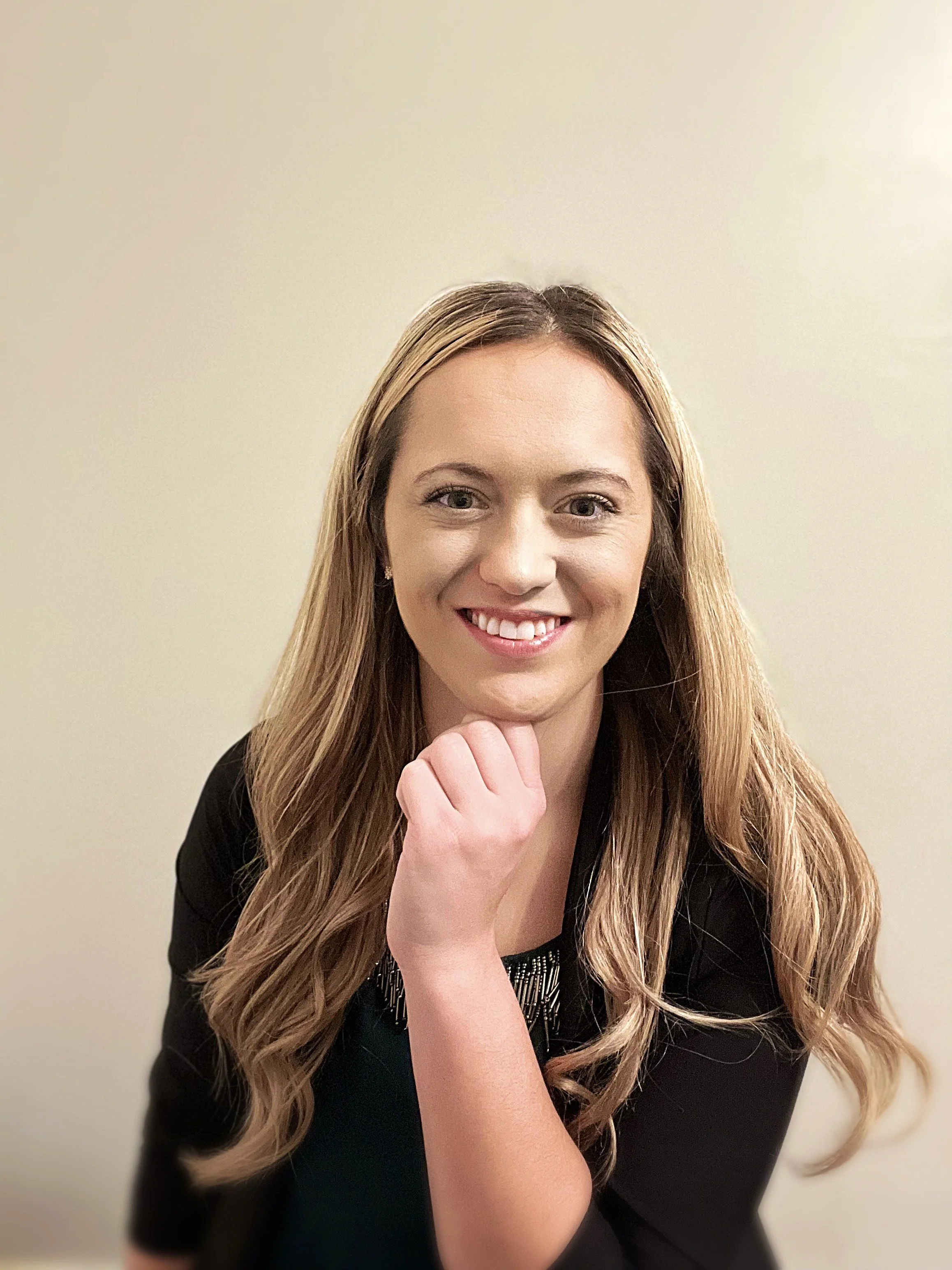 A woman with long wavy blonde hair smiling and holding her hand under her chin in front of a plain beige wall.