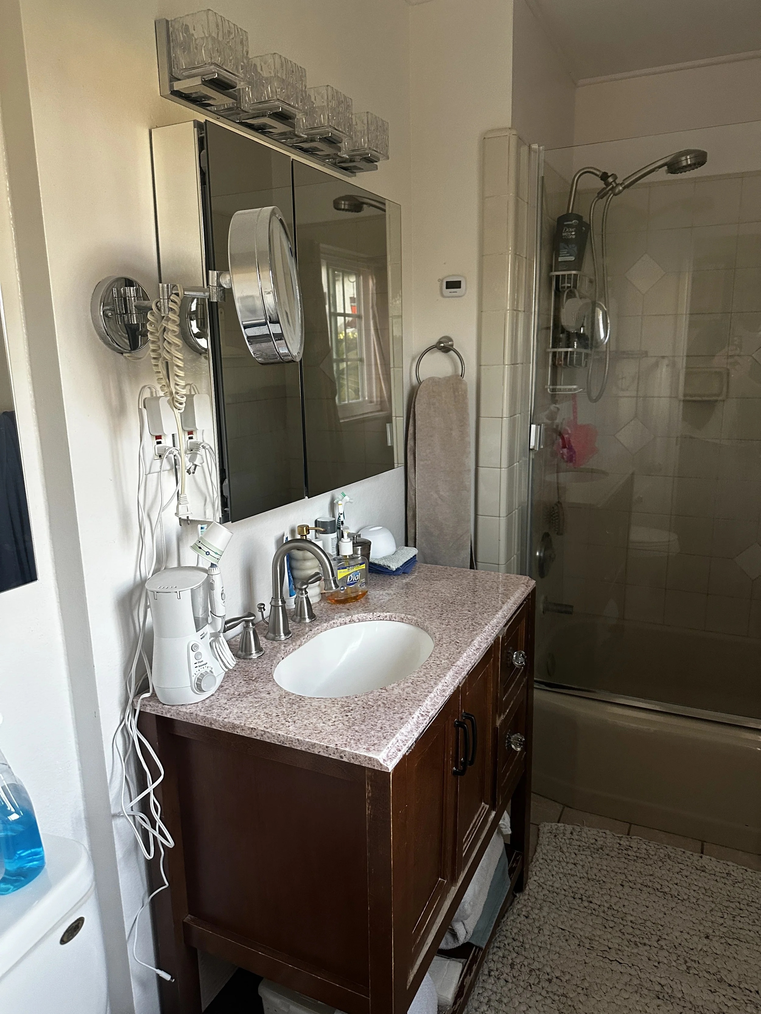 Bathroom with a granite countertop vanity with a sink, a mirror cabinet, soap dispenser, and electric dental device. Shower with glass door, showerhead, and toiletries.