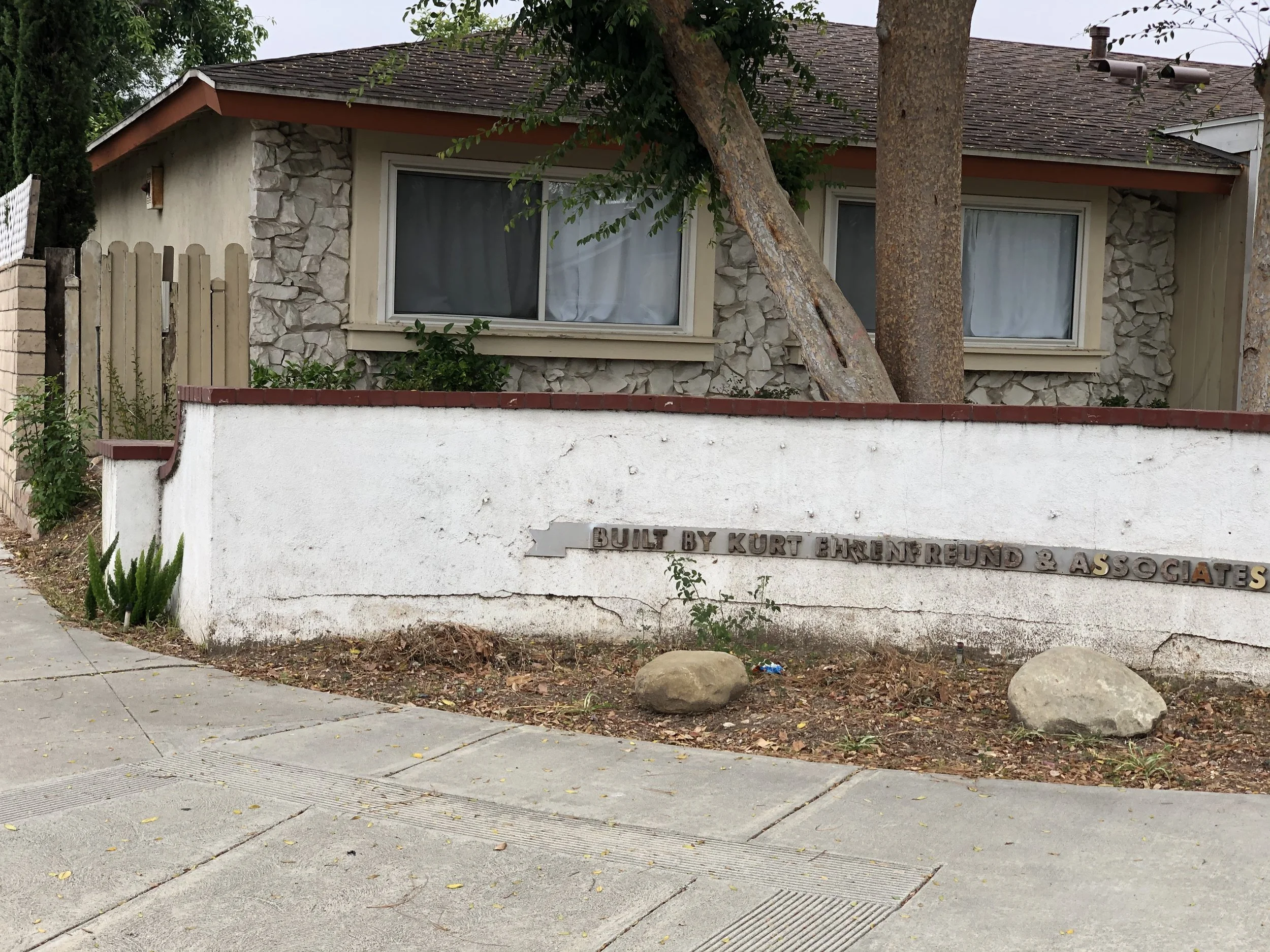 A house with beige stone walls, a sloped roof, and two large windows. There is a white wall in the foreground with a sign attached that reads 'Built by Kurt Ehrung & Associates.' Two large trees are in front of the house, partially obscuring it, and there is some greenery and dirt at the base of the house. The sidewalk and street are visible in the foreground.