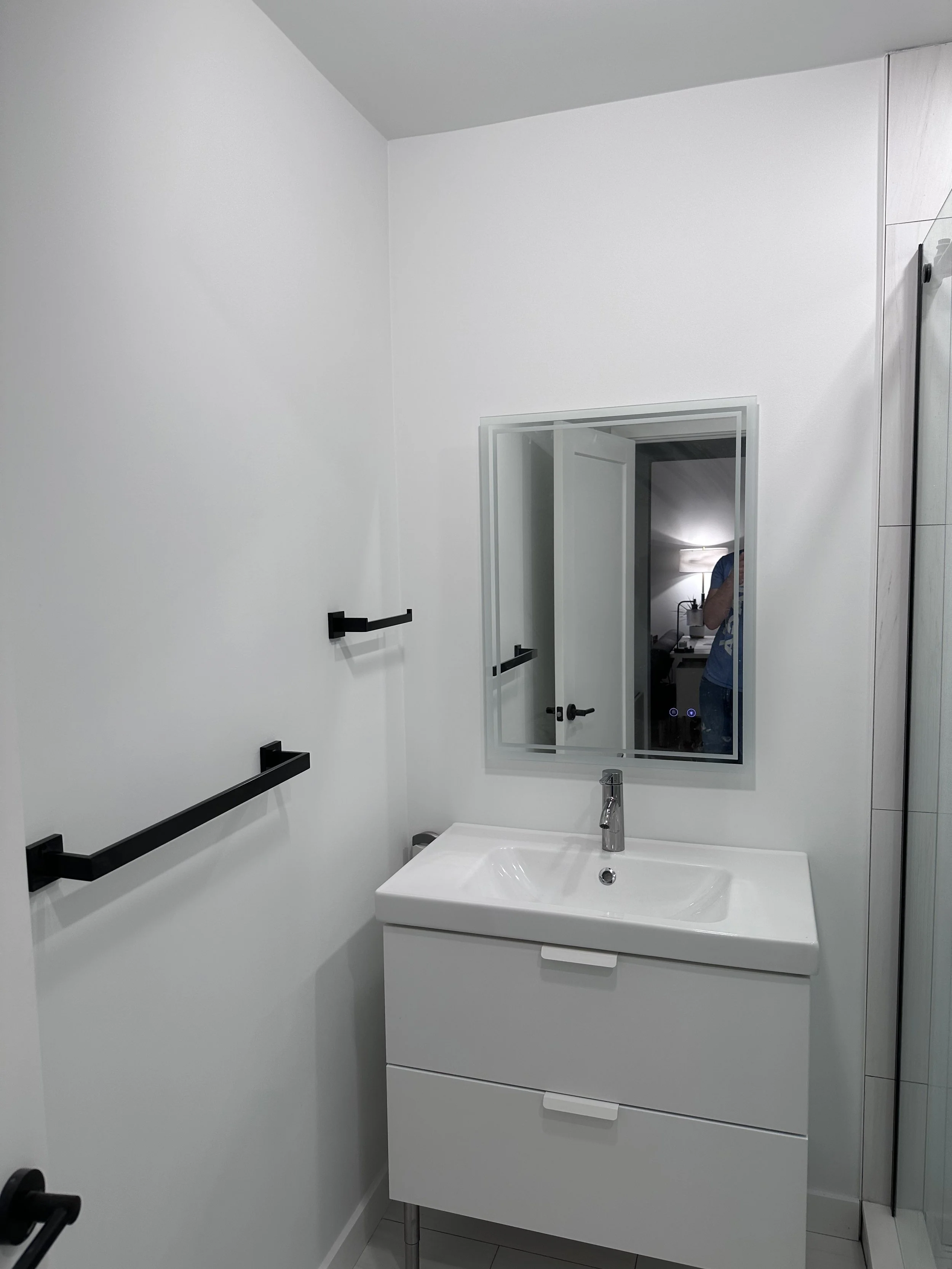 Modern bathroom with white sink vanity, mirror, black towel bars, and a door leading to a room with a lamp and a person taking the photo.