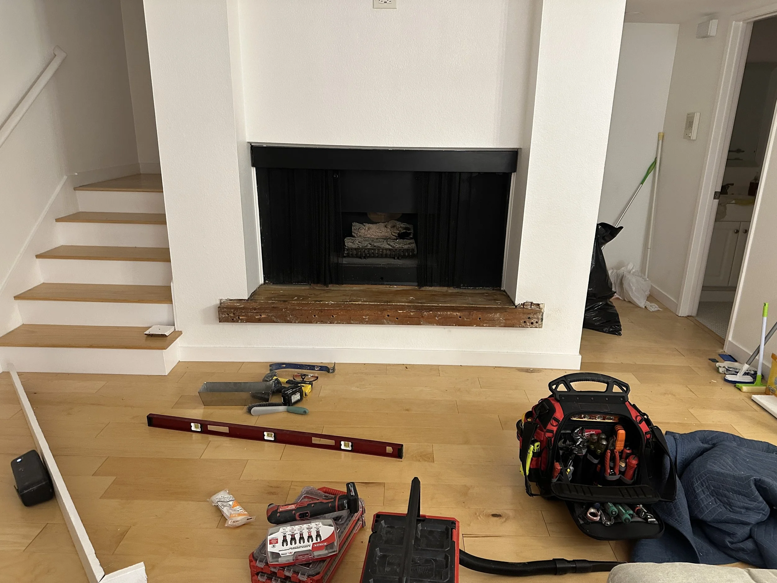 A room undergoing renovation with tools and supplies scattered on the floor including a level, a toolbox, a drill, and a level, with a black fireplace and an open doorway to a bathroom on the right, partially visible. The staircase on the left has wooden steps and a white railing.