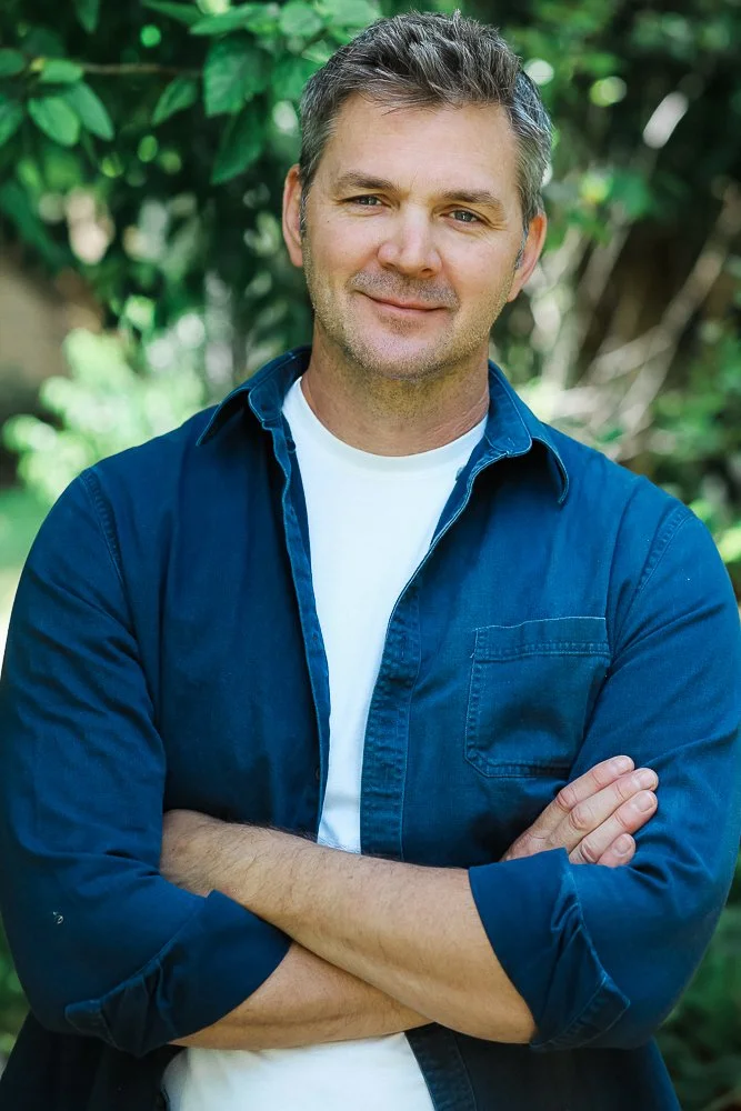 Man standing outdoors with arms crossed, wearing a dark blue button-up shirt over a white t-shirt, smiling slightly, with green foliage in the background.