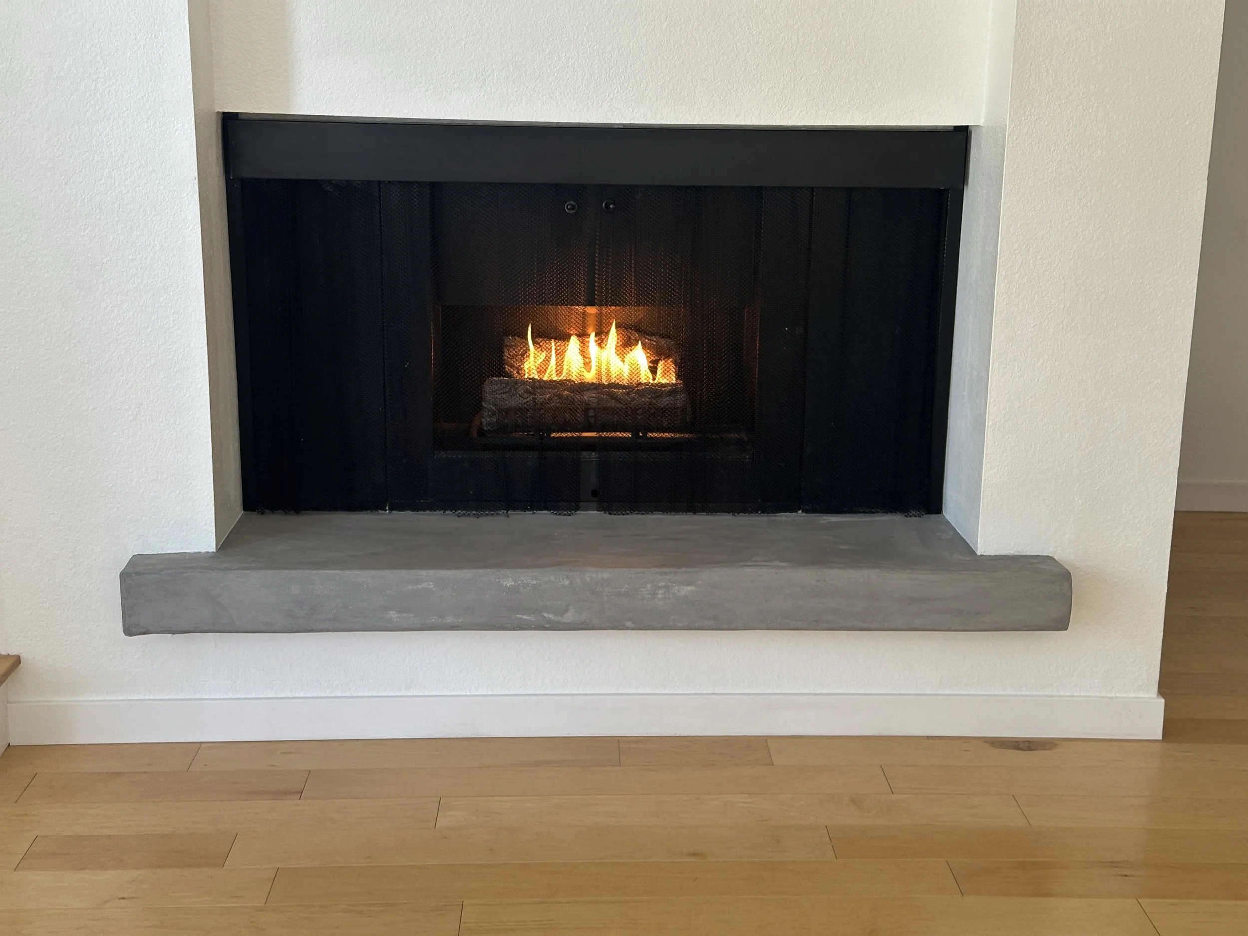 Modern fireplace with black metal frame and glass front, displaying a fire with glowing logs inside, built into a white wall with a concrete hearth and wooden floor.