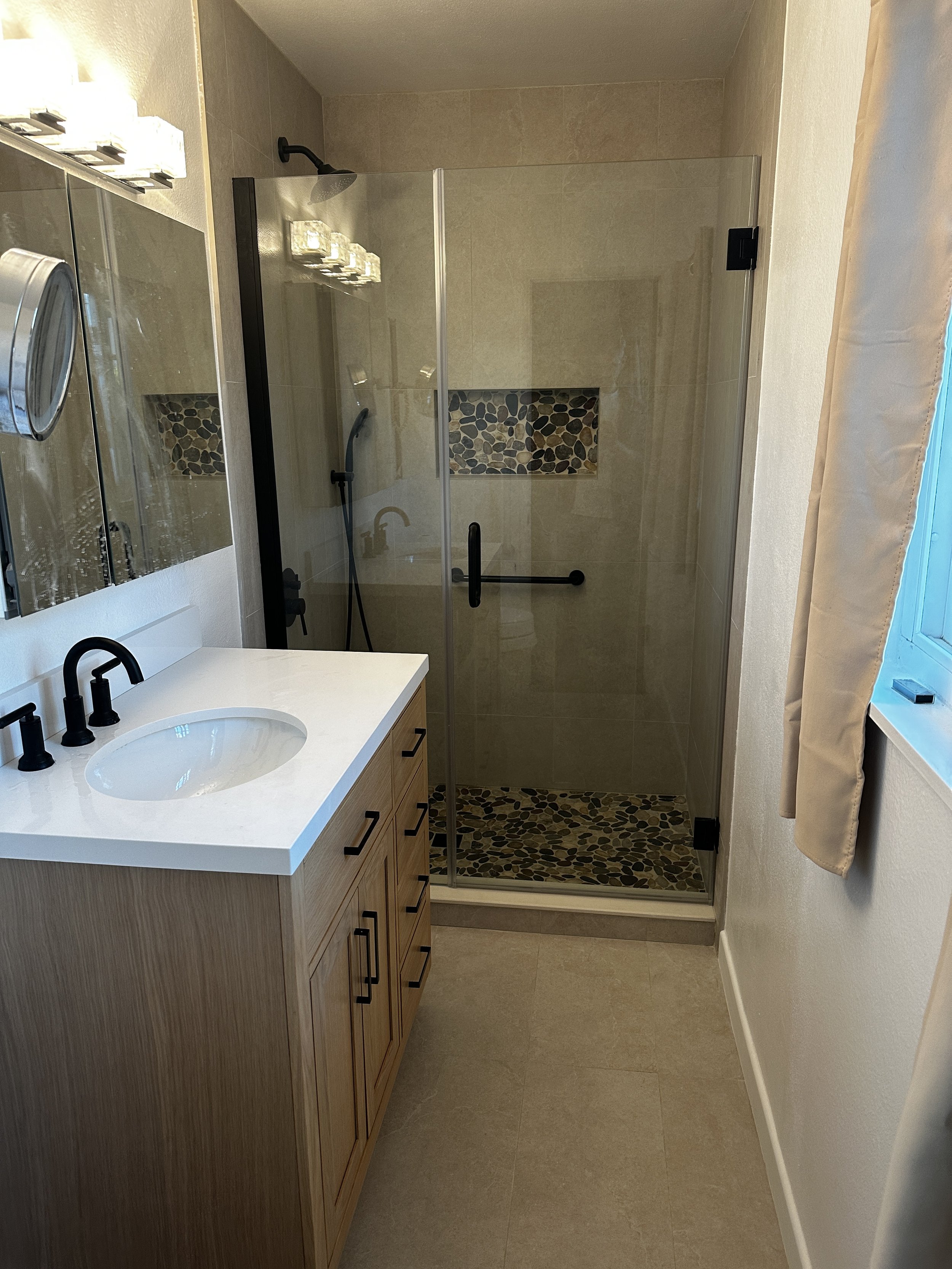 A modern bathroom with a wooden vanity, white countertop, black fixtures, and a walk-in shower with a glass door and pebble stone accents.