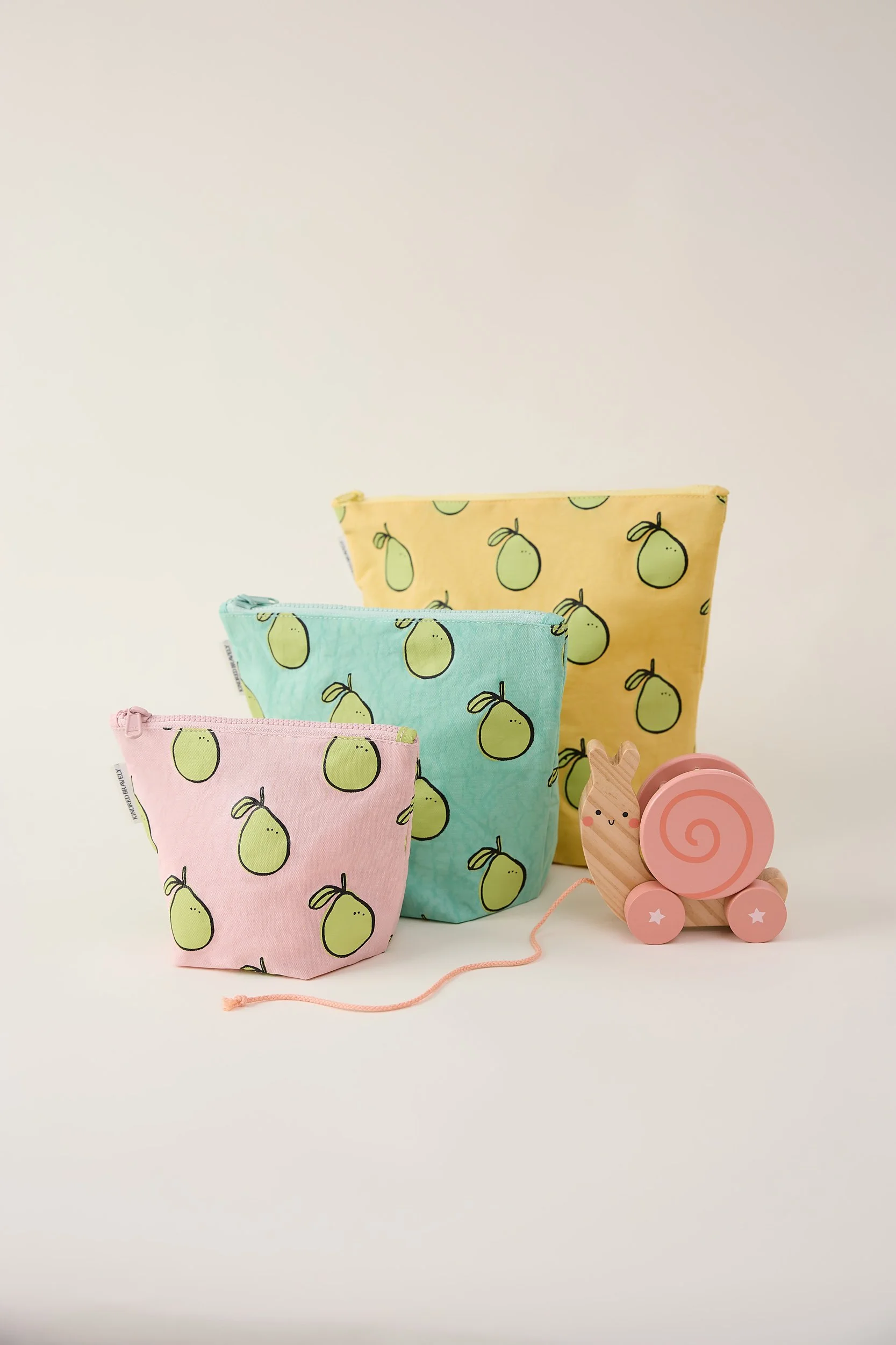 Retouched e-commerce shot of three pastel-colored pouches with pear patterns, a wooden toy unicorn, and a pink snail toy.