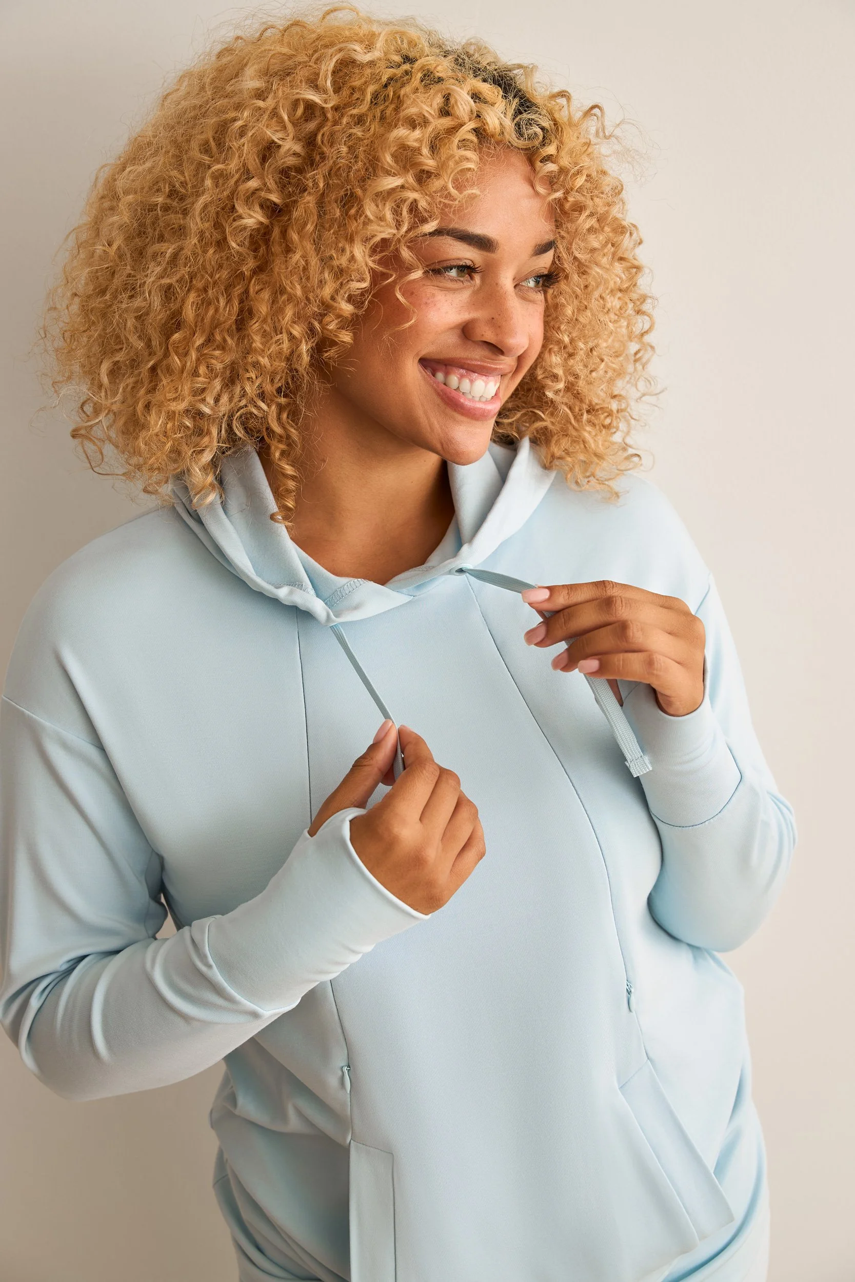 A retouched e-commerce shot of a woman with curly blond hair wearing a light blue hoodie, smiling, holding the strings of the hoodie. Some wrinkles have been removed from the clothing and the color has been adjusted.