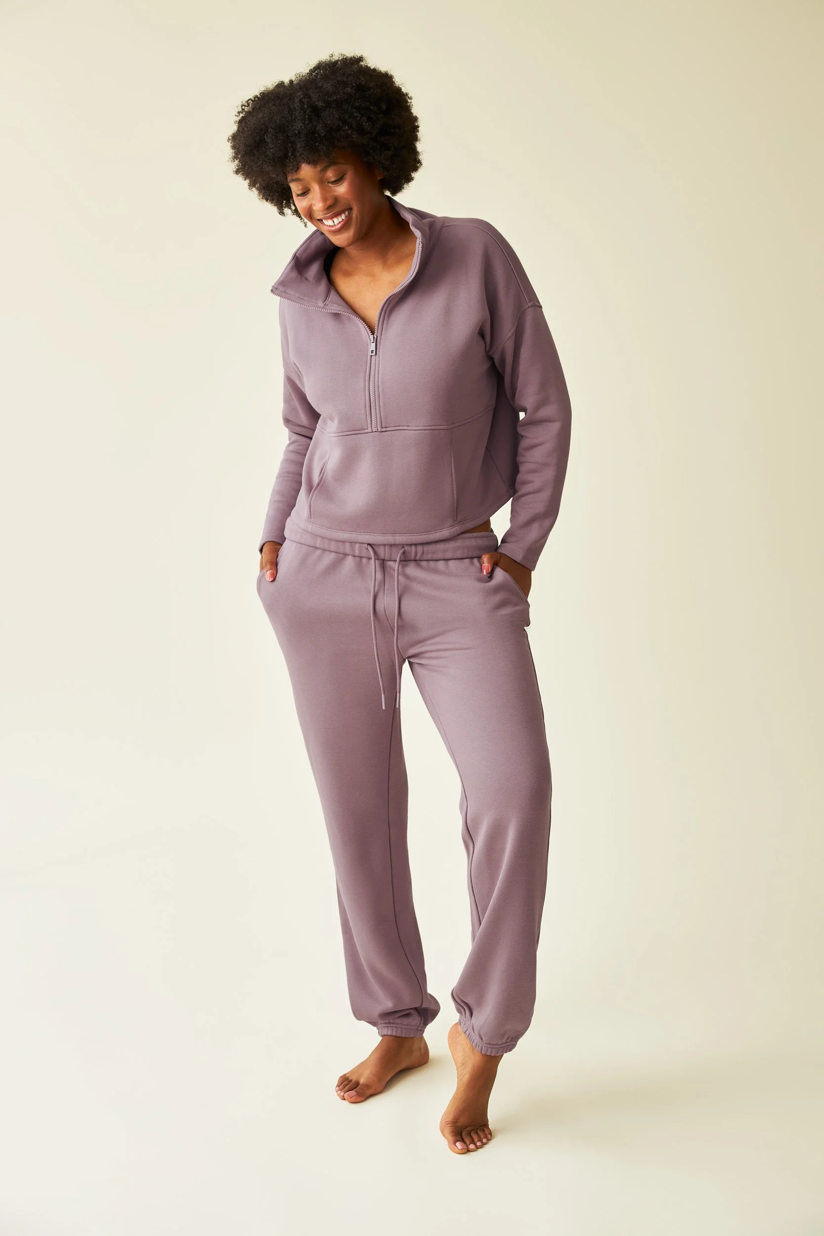 Retouched e-commerce shot of a model in mauve loungewear standing barefoot against a plain light background, smiling and looking down. Wrinkles have been removed from clothing and product has been color corrected.