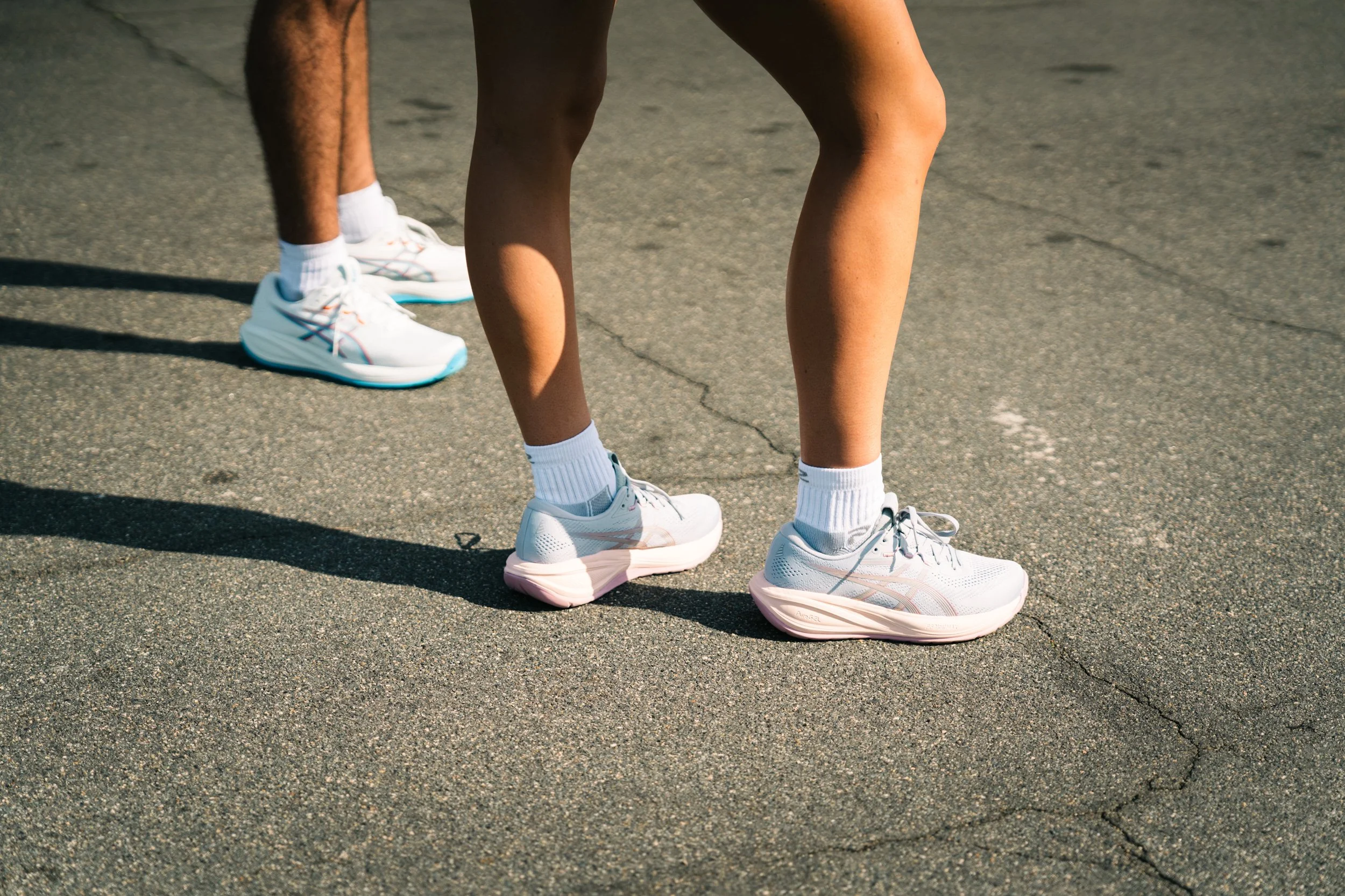 Unretouched close-up of two people wearing white Asics shoes and white socks, standing on a paved outdoor surface, with shadows cast on the ground.