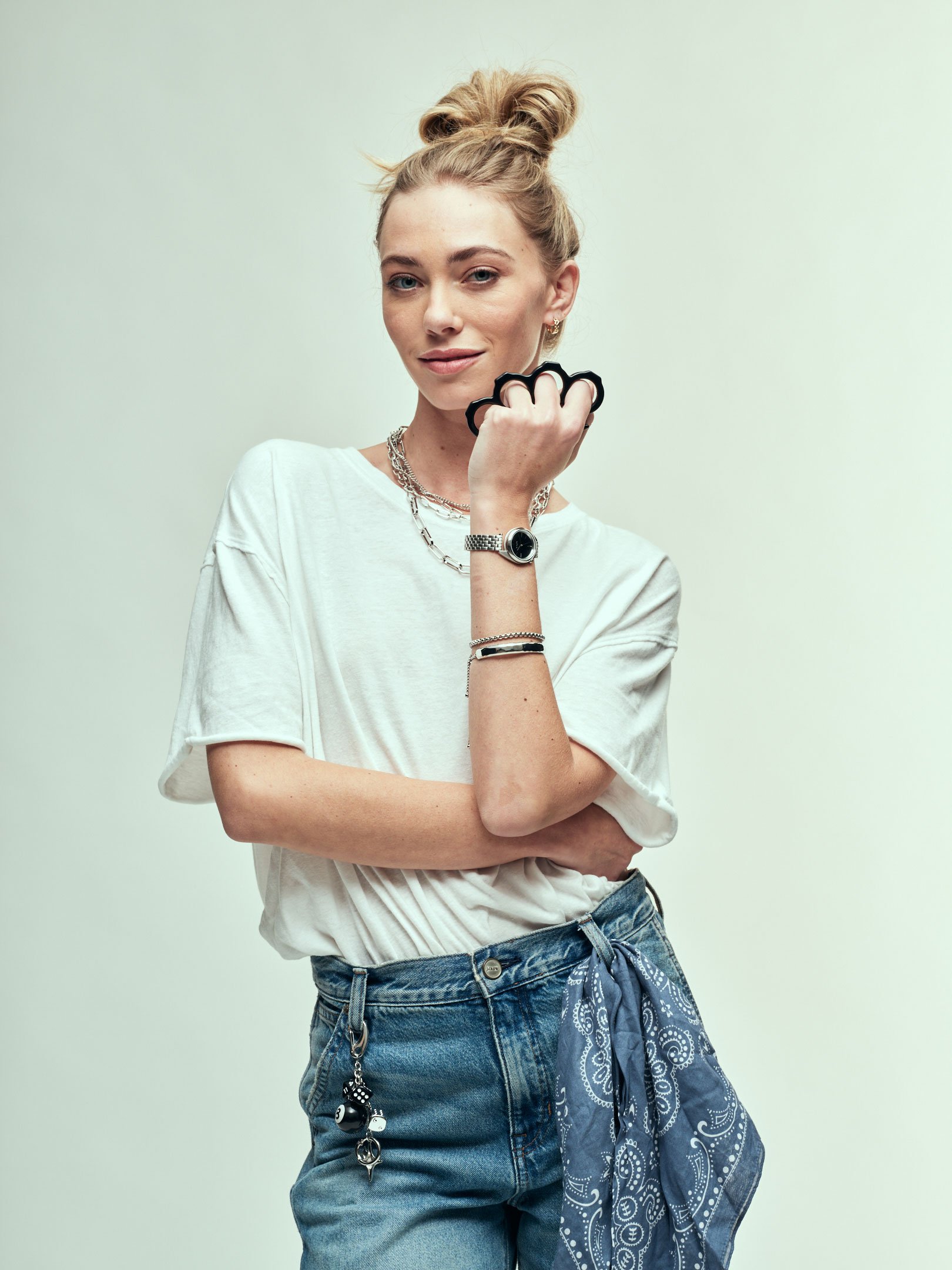 Original image of a young woman with blonde hair in a messy bun, wearing a loose white t-shirt with rolled sleeves, blue jeans, layered jewelry, wristwatch, and a blue patterned bandana tied around her waist, standing against a plain background.