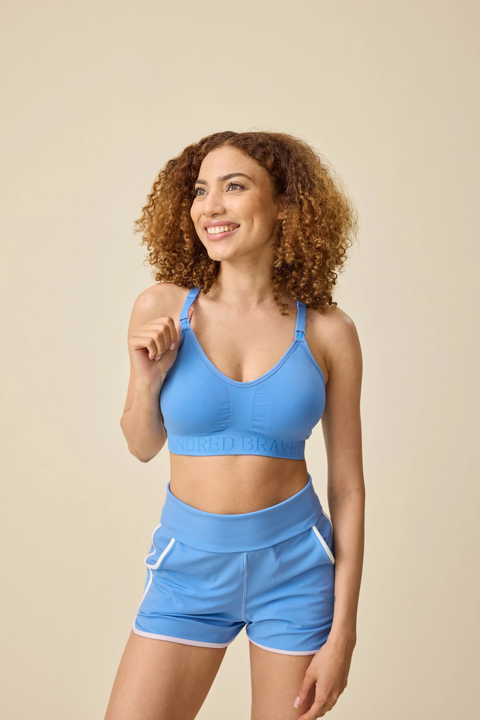 Original image of a smiling woman with curly reddish hair wearing a blue sports bra and matching shorts, standing against a plain beige background