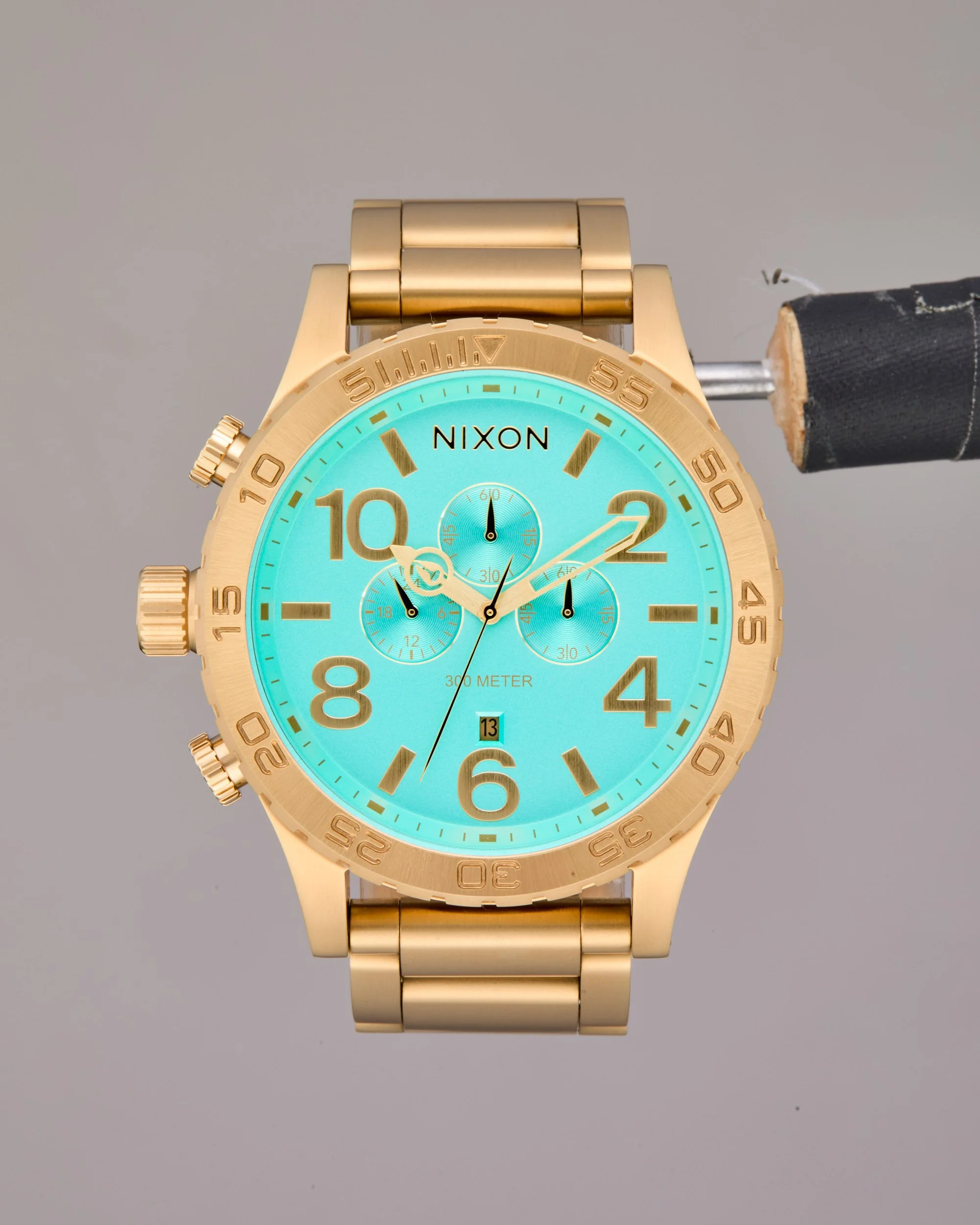 Original unretouched e-commerce image of a gold wristwatch with a turquoise dial, featuring chronograph subdials and gold markers, with the 'NIXON' on the face.