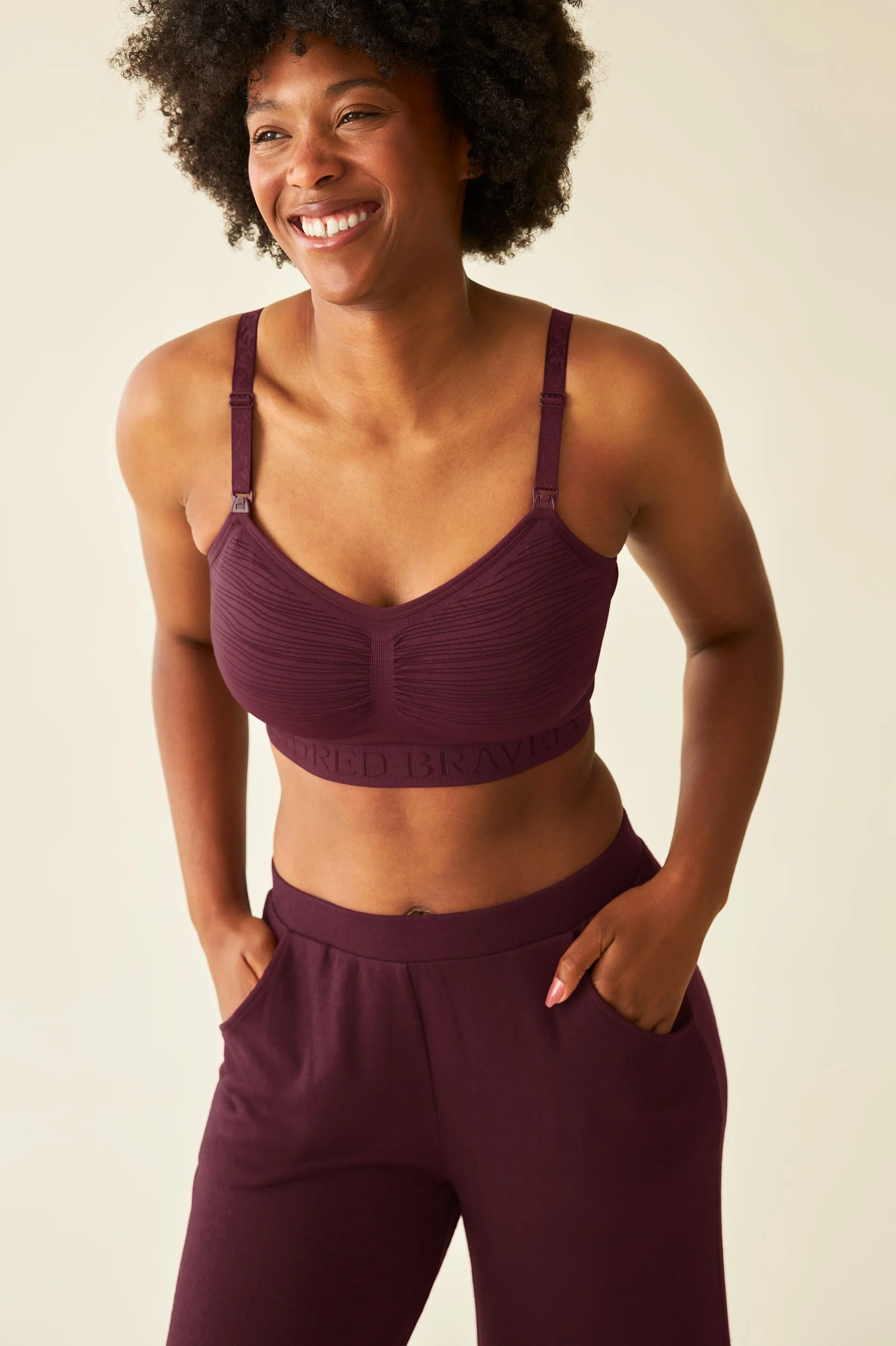 Retouched shot of a model with curly hair smiling and wearing a maroon sports bra and matching pants, with her right hand in her pocket. Color has been corrected and wrinkles removed from clothing.