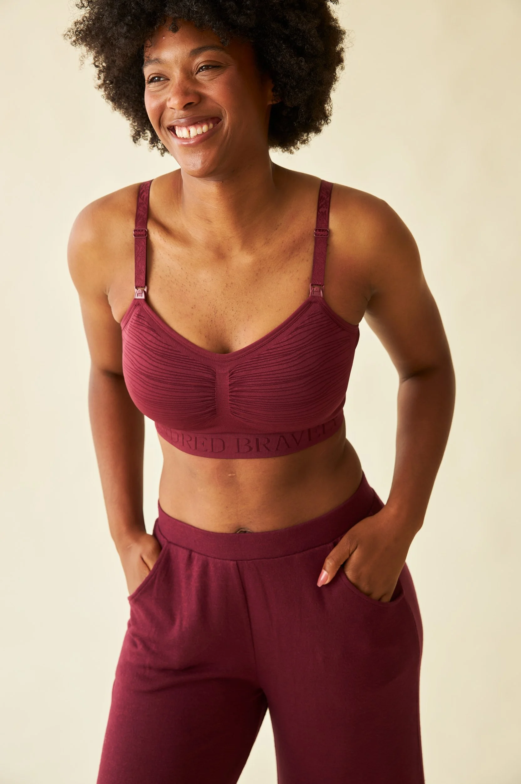 An unretouched shot of a model with curly hair smiling and wearing a maroon sports bra and matching pants, with her right hand in her pocket