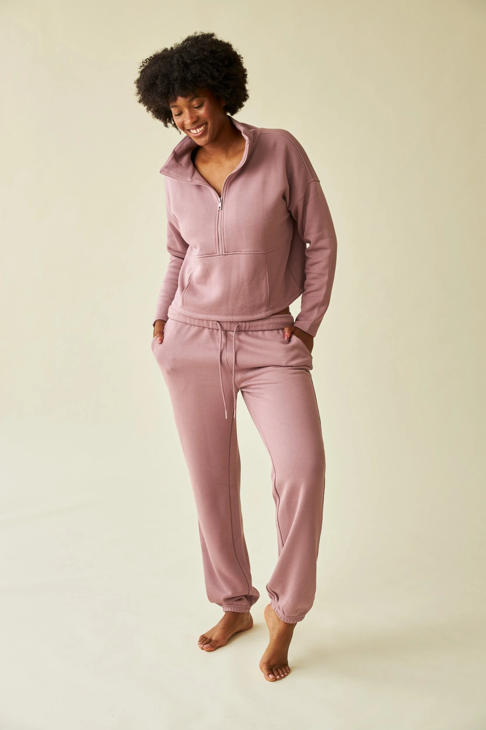 Unretouched e-commerce shot of a model in mauve loungewear standing barefoot against a plain light background, smiling and looking down. Wrinkles have been removed from clothing and product has been color corrected.