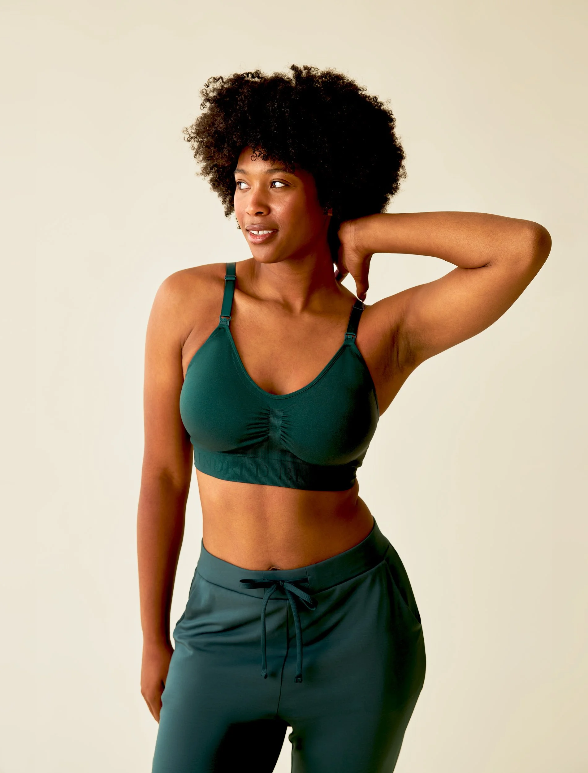 A retouched e-commerce shot of a model with curly hair wearing a teal sports bra and matching joggers, touching the back of her neck with her right hand. Wrinkles have been smoothed and colors have been corrected.