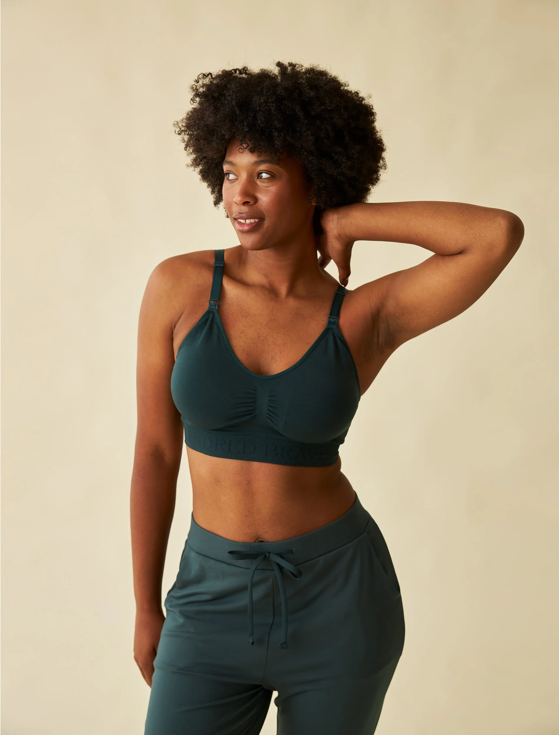 An unretouched e-commerce shot of a model with curly hair wearing a teal sports bra and matching joggers, touching the back of her neck with her right hand. Wrinkles have been smoothed and colors have been corrected.