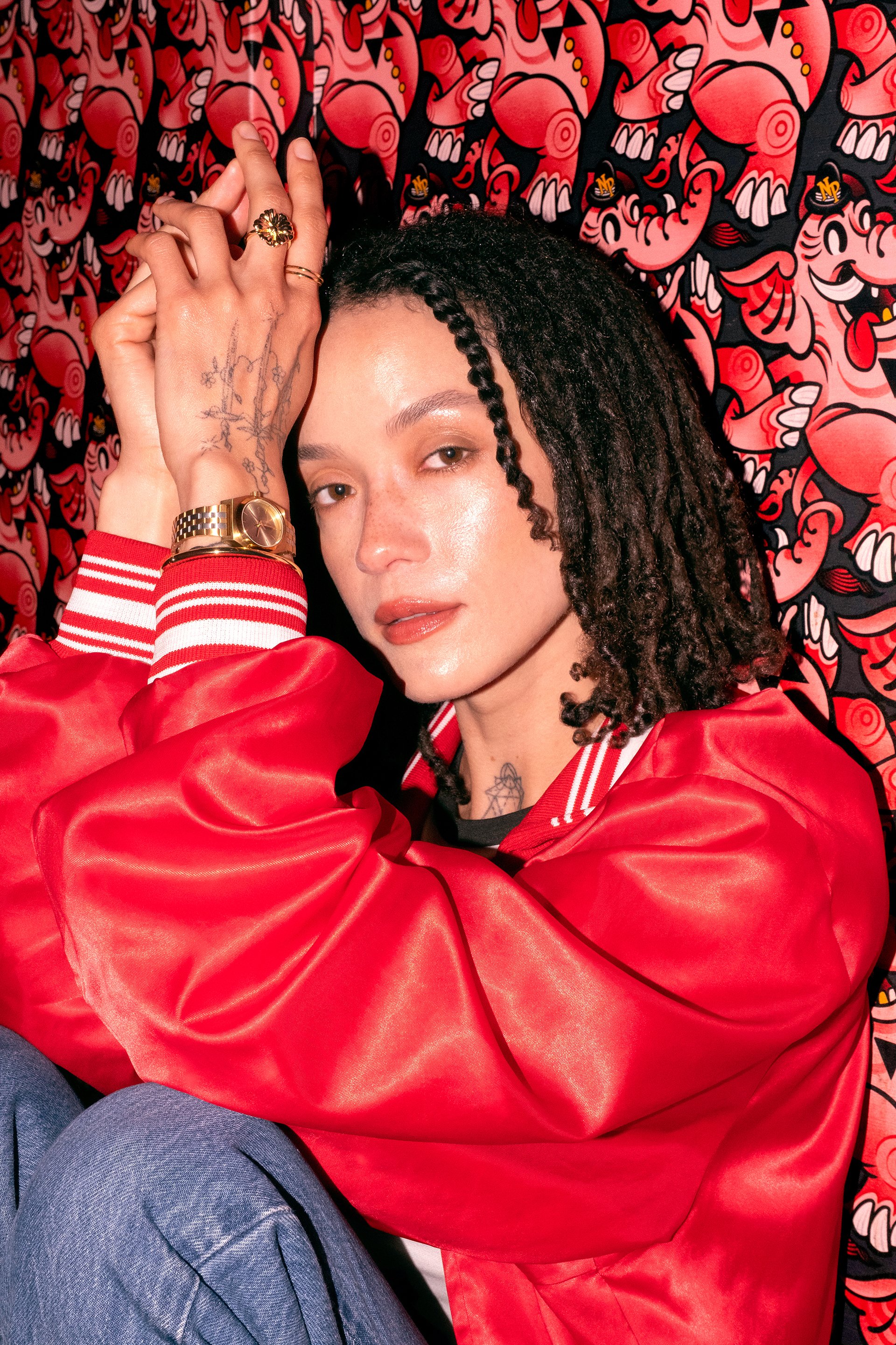 Retouched photo of a woman with dark curly hair, wearing a red satin jacket, resting her hand with rings and a gold watch on her forehead against a vibrant red and black cartoon patterned background.