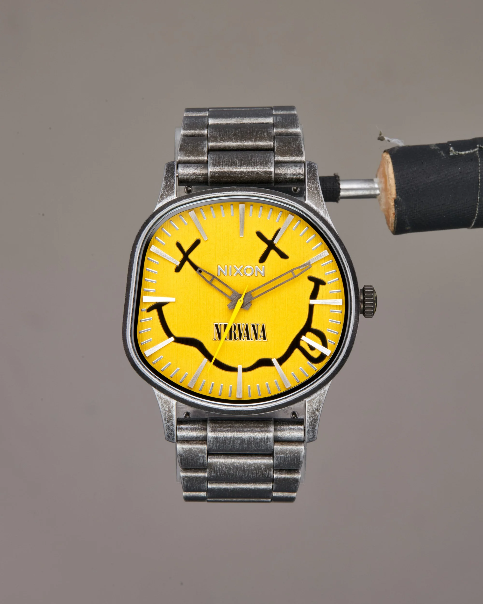 Original shot of the NIRVANA Wobble wristwatch with a yellow face, black hour markers, and a black smiley face design, featuring the text 'NIXON' and 'NIRVANA', on a metal band.