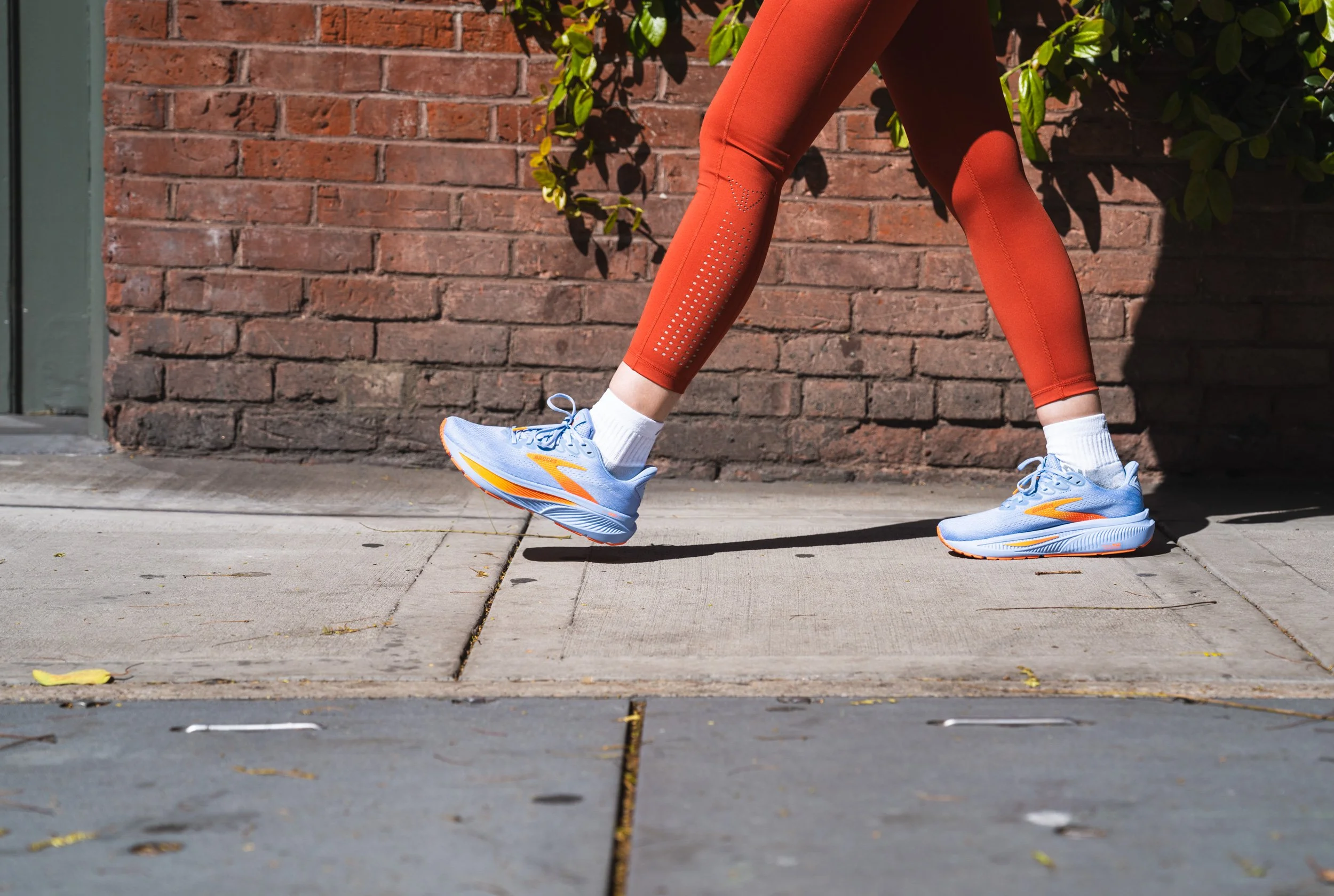 Person walking on sidewalk wearing orange athletic leggings and blue running shoes with orange accents, against a brick wall and green foliage background.
