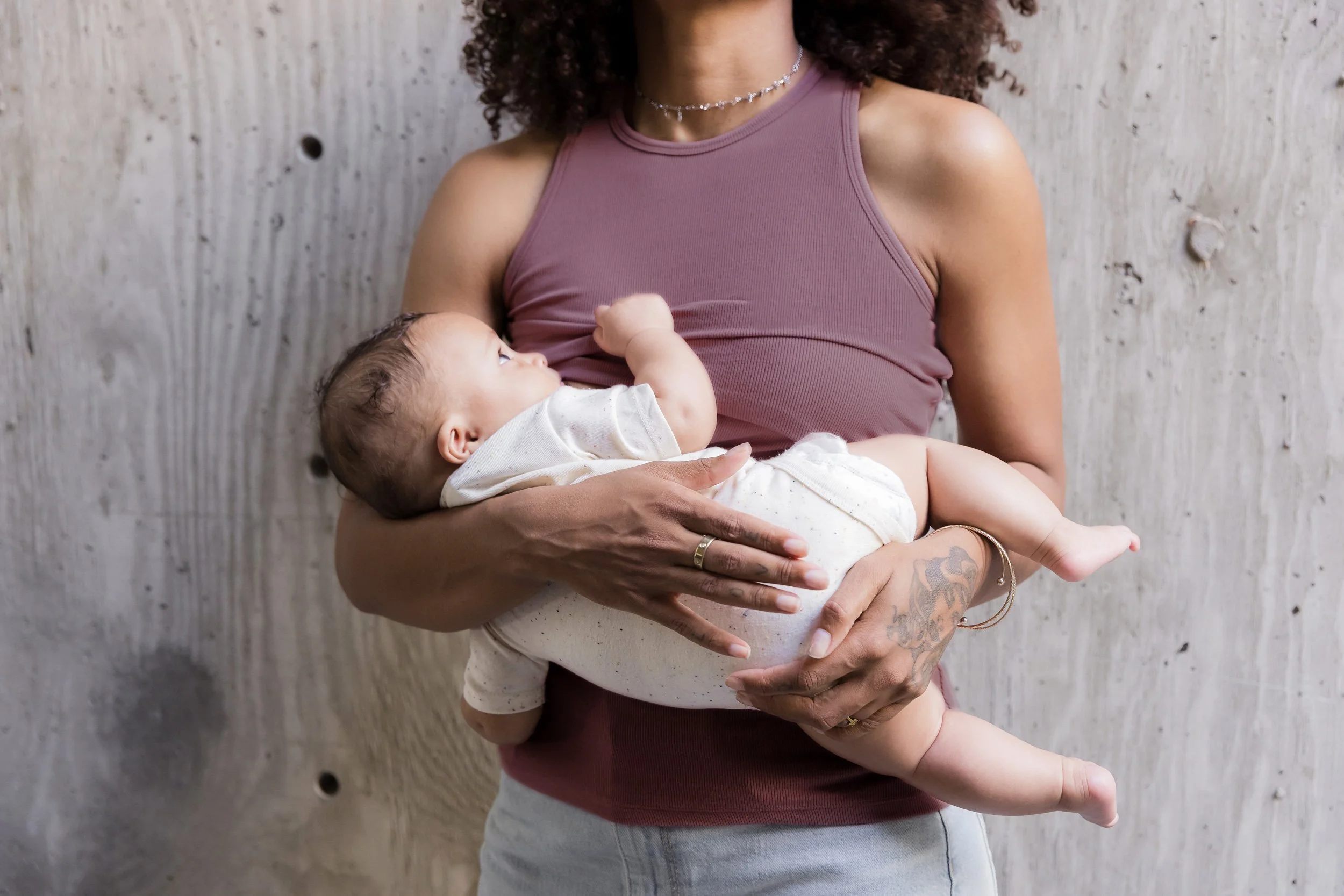 Retouched image of a woman holding a baby against a concrete wall. The woman is wearing a mauve sleeveless top and a silver necklace. The baby is dressed in a white outfit and is looking up at the woman.
