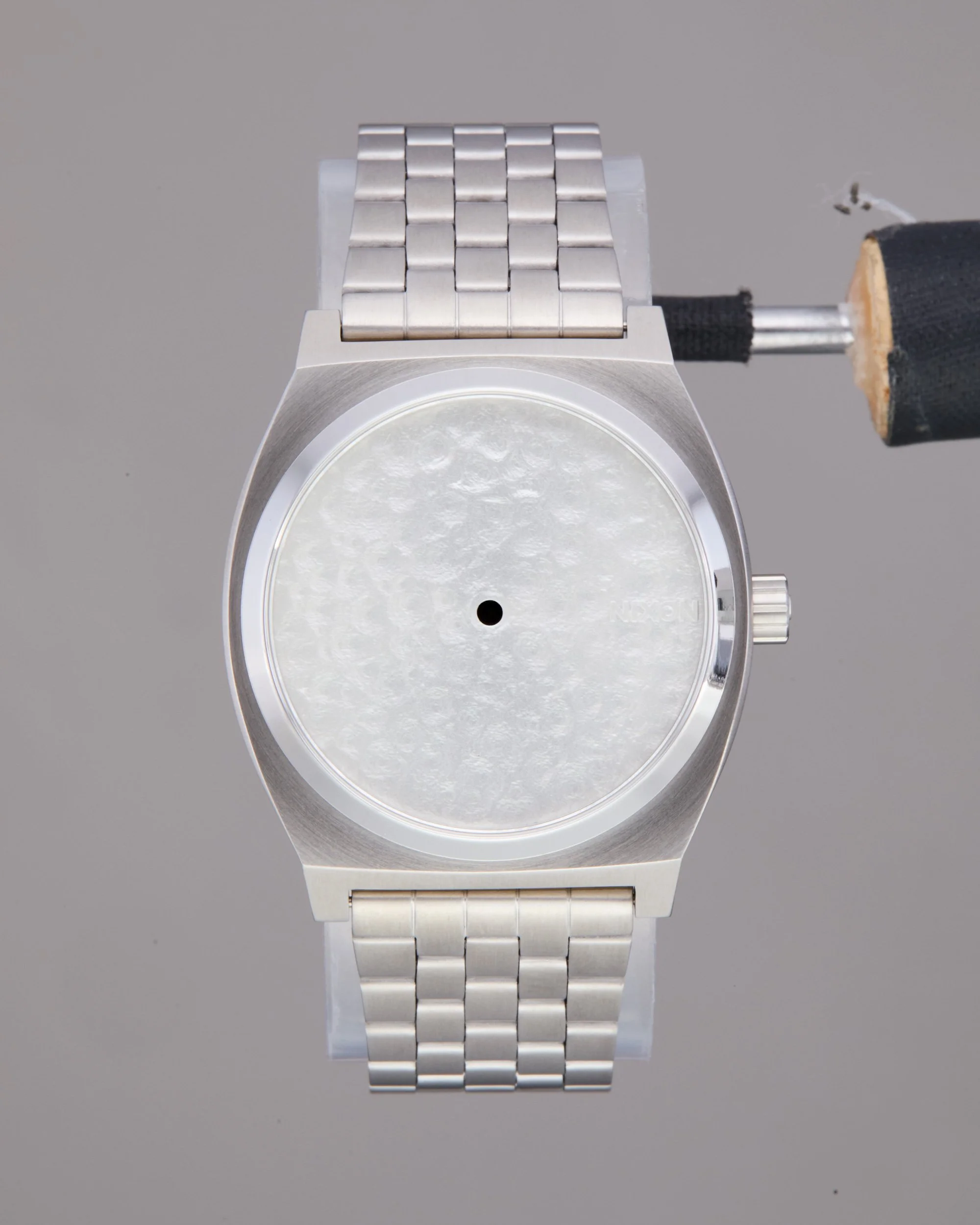 Original shot of a silver wristwatch with a metal band and a round white face, positioned against a plain gray background. It is missing the indices, hands, and logo.