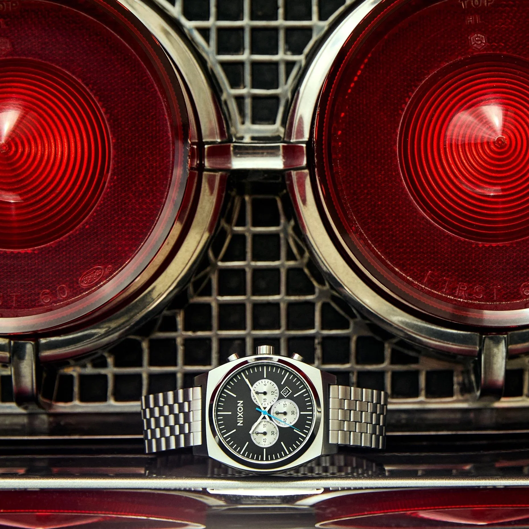 Close-up of a black Nixon wristwatch with silver bracelet, displaying the time, positioned in front of two red car tail lights on a chrome grid surface.