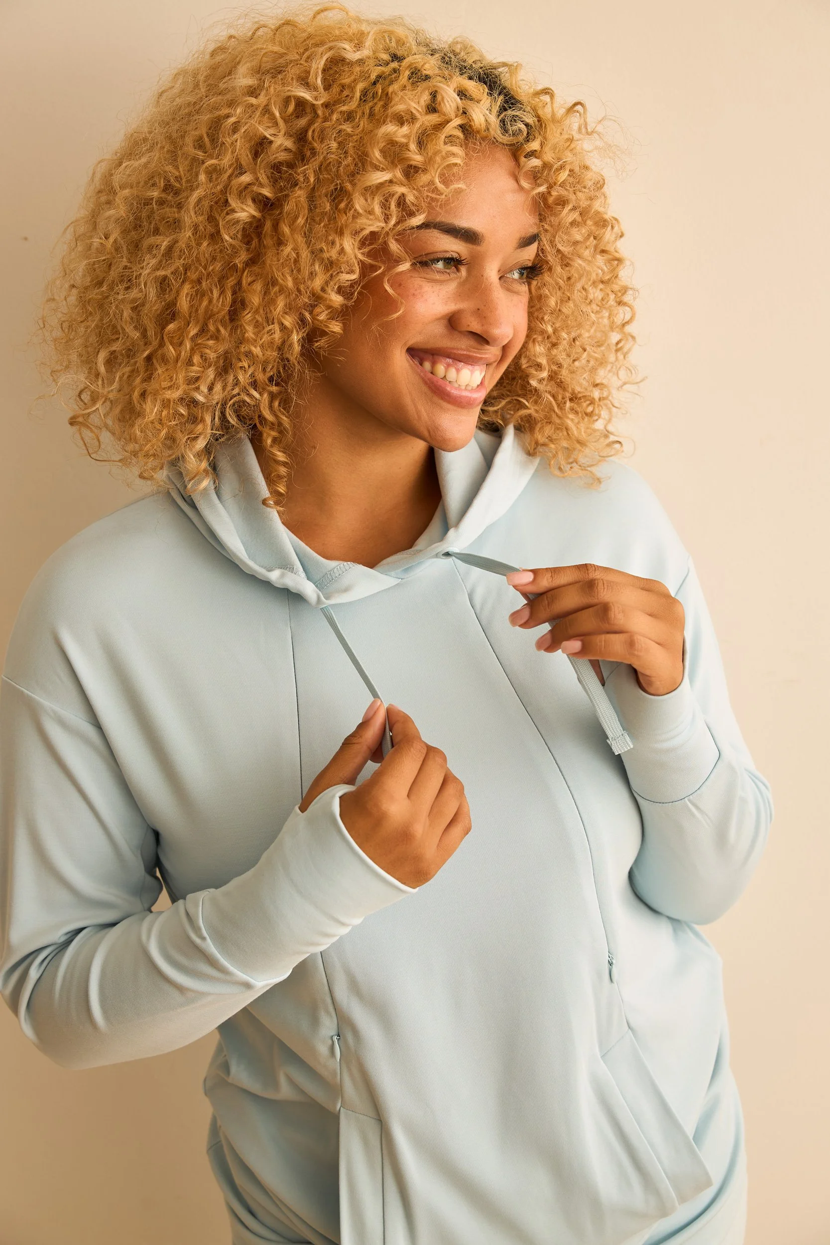 An un retouched e-commerce shot of a woman with curly blond hair wearing a light blue hoodie, smiling, holding the strings of the hoodie. 
