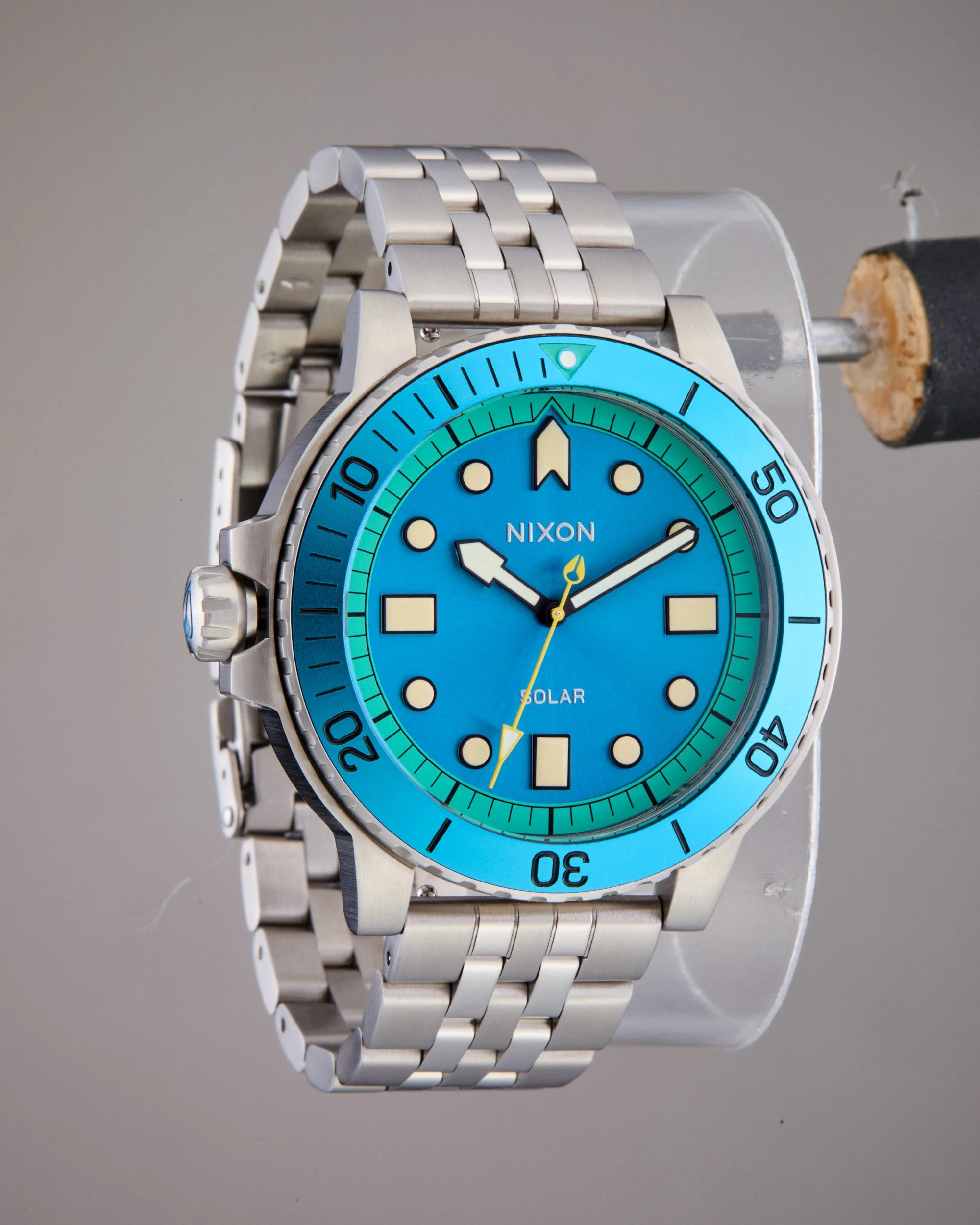 Original shot of a Nixon solar-powered wristwatch with a blue dial, luminous hands, and a metal band, displaying time against a plain background.