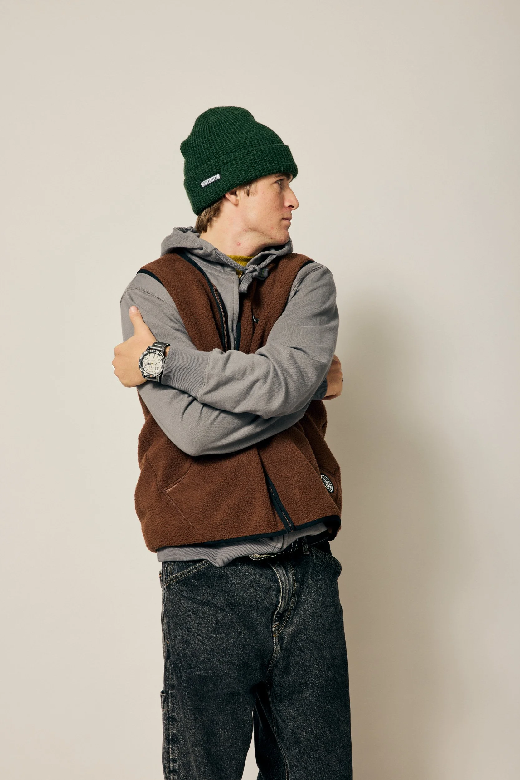 Original e-commerce shot of a young man with brown hair wearing a green knit beanie, a gray hoodie under a brown fleece vest, dark jeans, and a watch, standing against a plain beige wall, looking to his right with arms crossed.