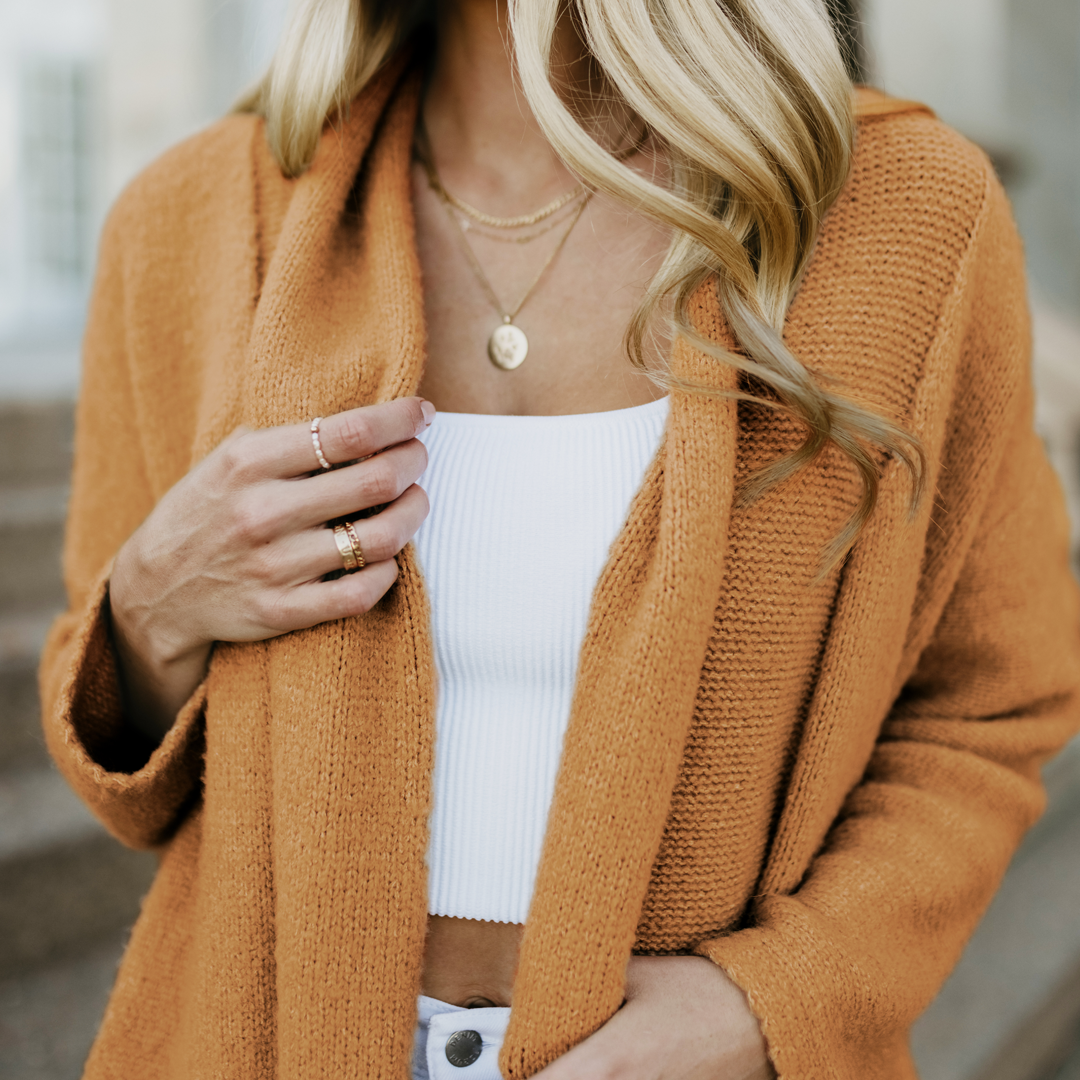 Retouched and color swap done to a shot of a woman shown from her shoulders to her waist, wearing an orange cardigan over a white top, with layered necklaces and rings on her fingers.