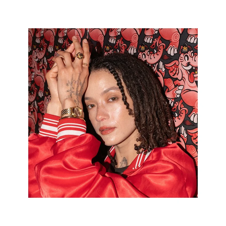 Original image of a woman with curly, styled hair and tattoos, wearing a red jacket, jewelry, and makeup, posing with her hand on her head against a patterned background.