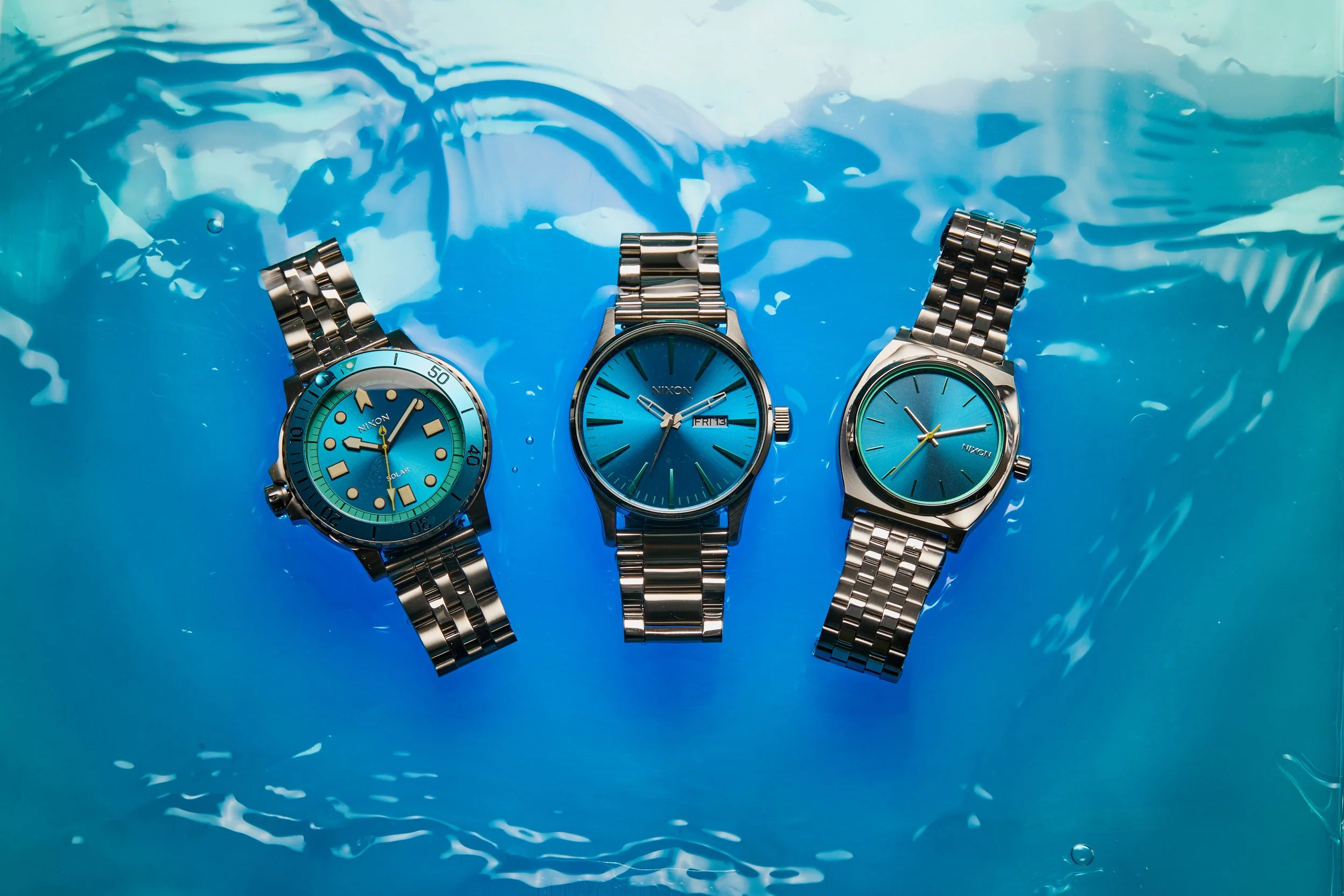 Unretouched image of three metal wristwatches submerged in clear blue water with ripples and reflections.
