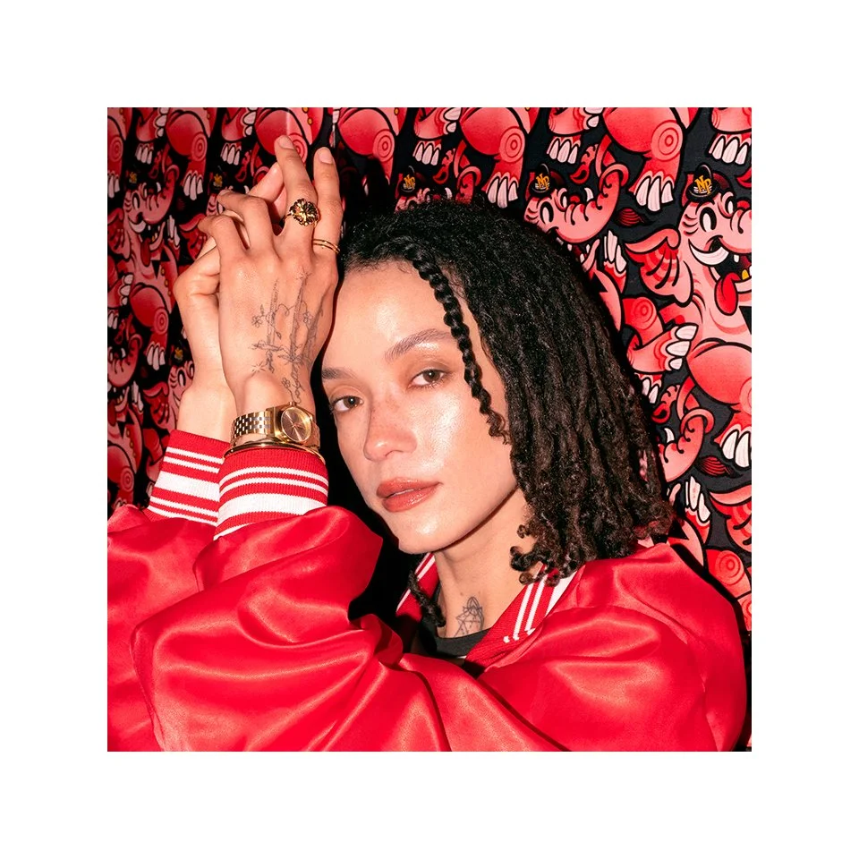 Retouched photo of a woman with curly black hair and tattoos on her hand and neck, wearing a red jacket with white striped cuffs, posing with her hands near her face against a red and black abstract patterned background.