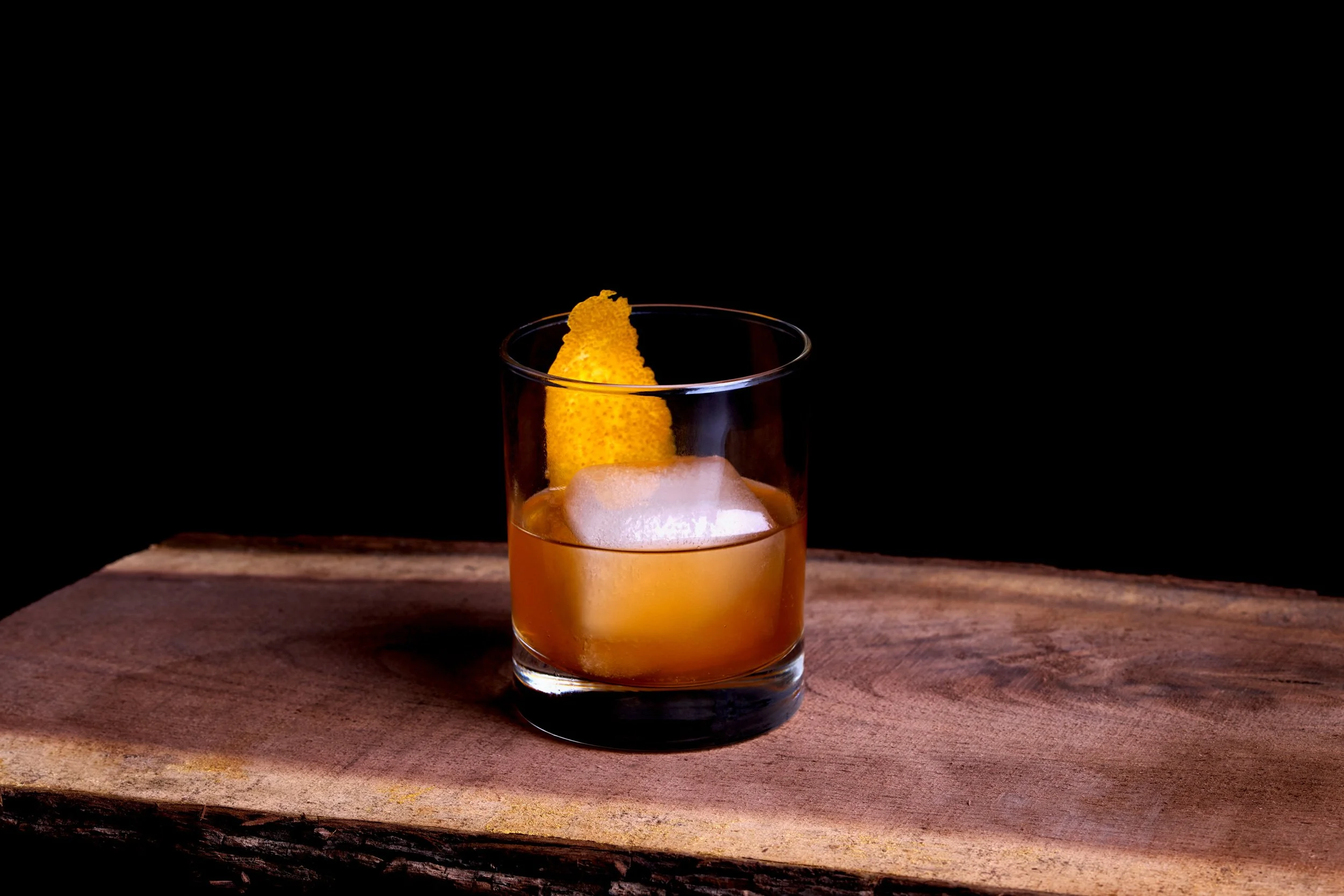 Retouched shot of a glass of whiskey with a lemon peel garnish on a wooden surface against a black background.