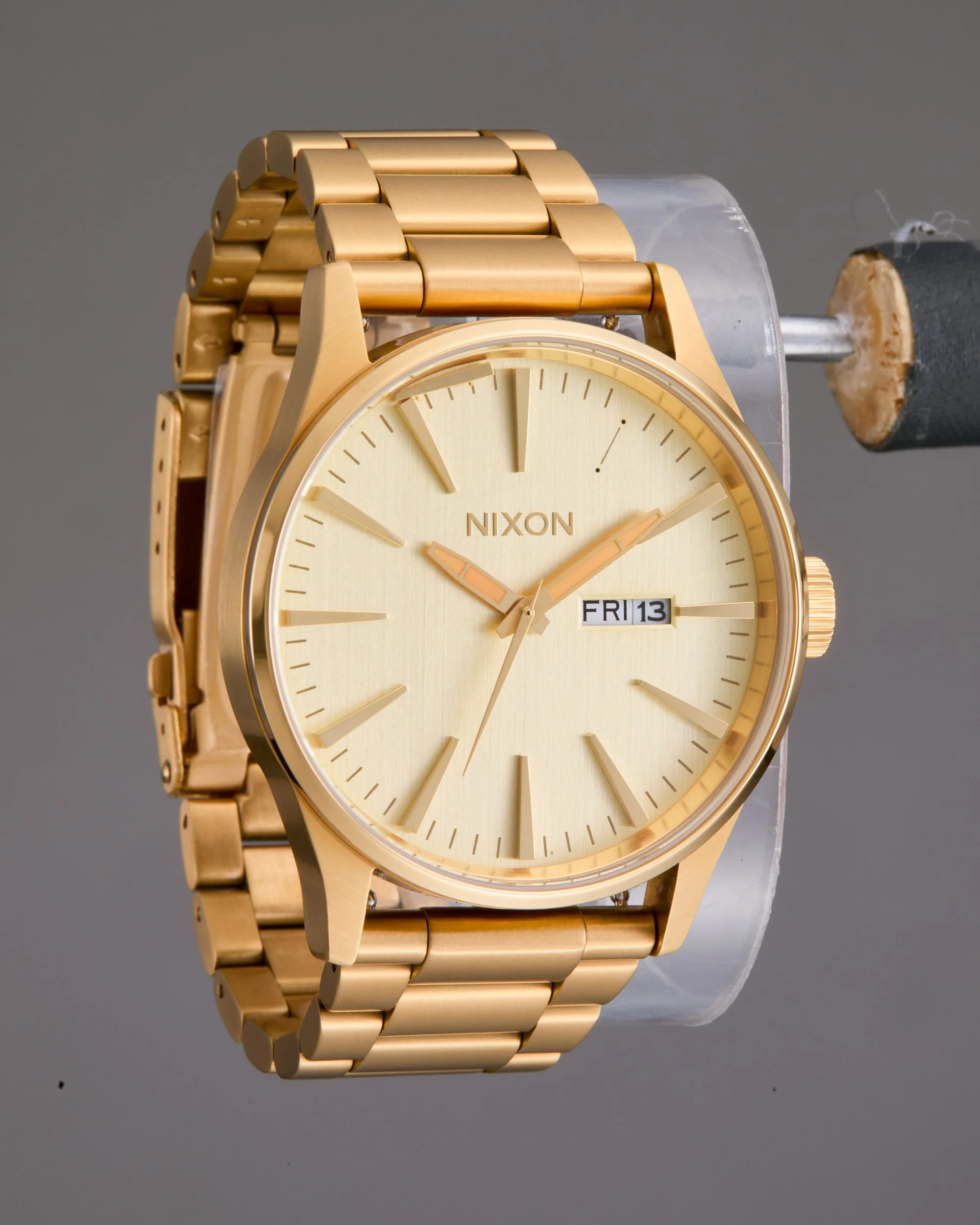 Original unretouched shot of a gold-colored Nixon wristwatch with a metal band, displayed on a stand with a gray background.