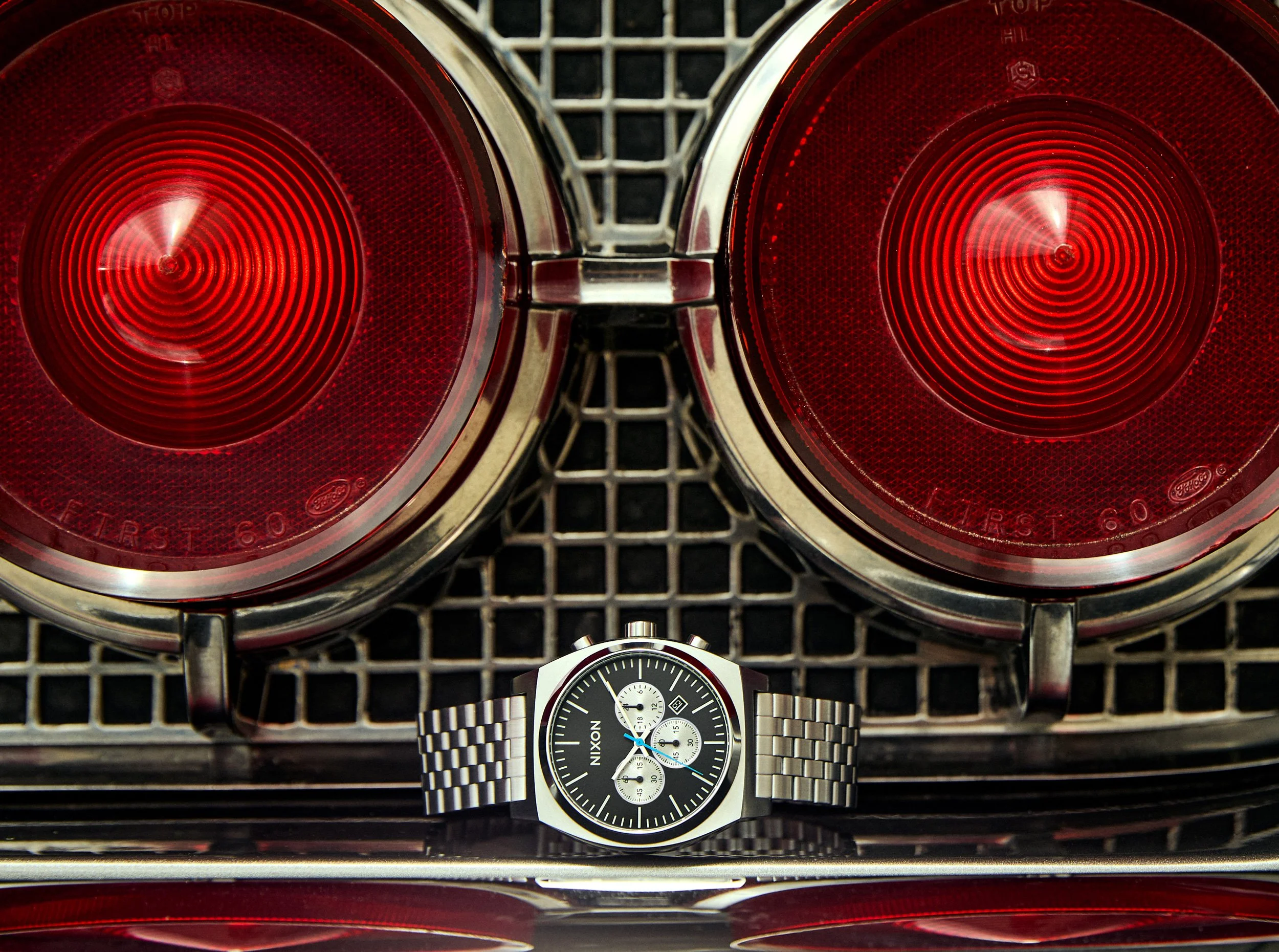 Close-up of two red car taillights on a vehicle bumper with a black and silver wristwatch with a metal band placed in front of them on a black metal grate surface.