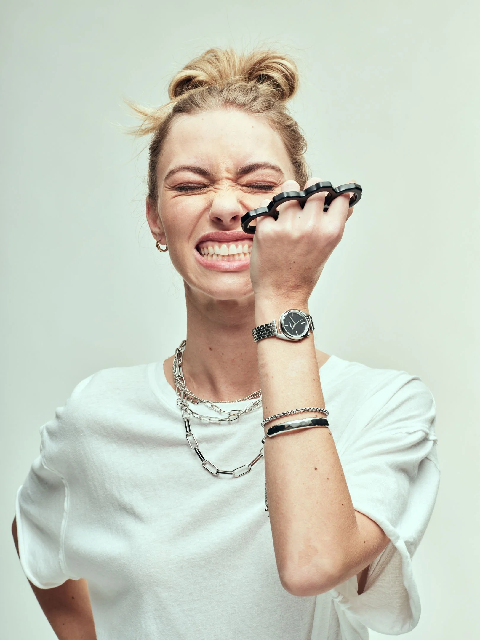 Original shot of a woman with blonde hair in a bun, wearing a white shirt and silver jewelry, is covering her face with a black brass knuckles, making a facial expression with her eyes closed and teeth showing.