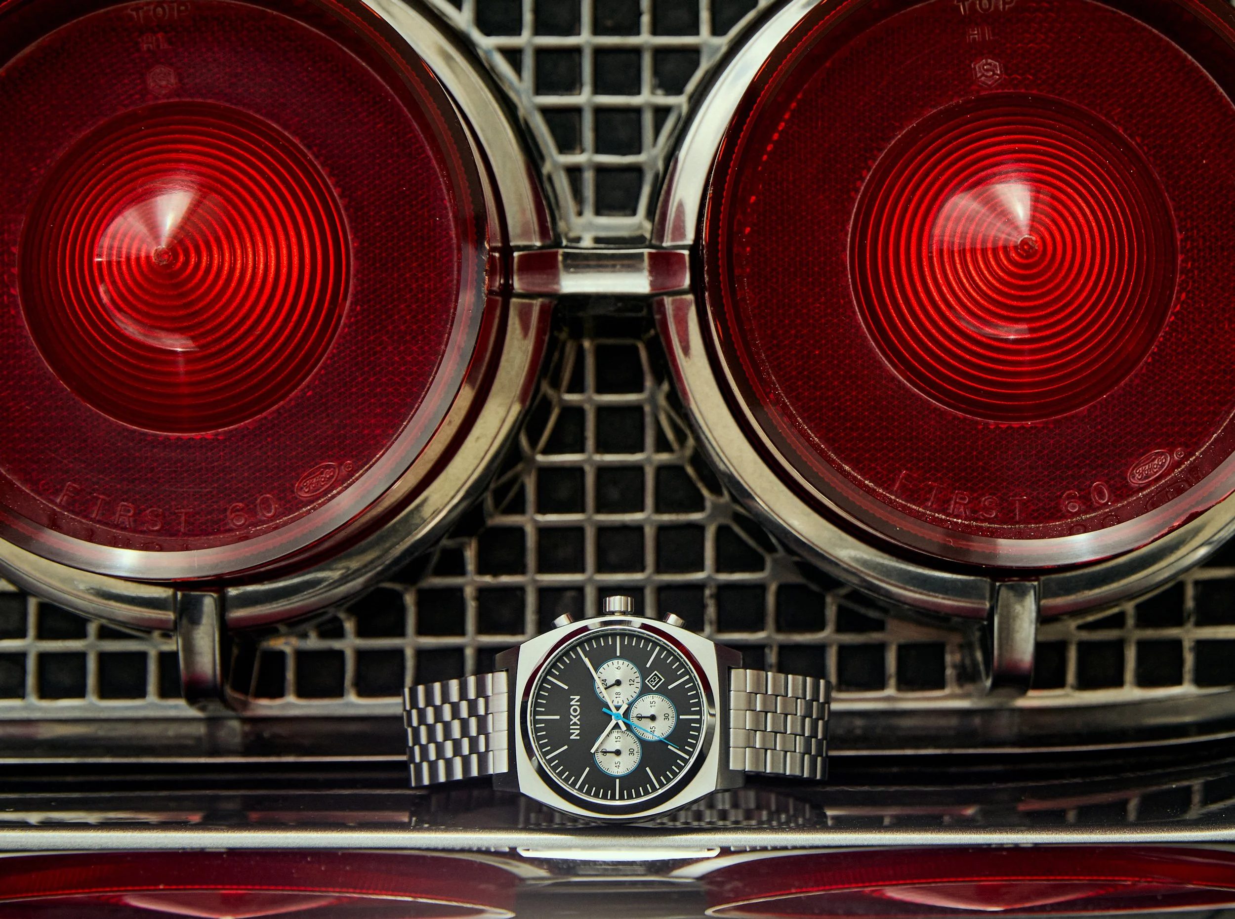 Close-up of two red round car taillights on a vehicle, with a silver wristwatch with a black dial and silver bracelet placed in front of them. The background is a metallic grid surface.