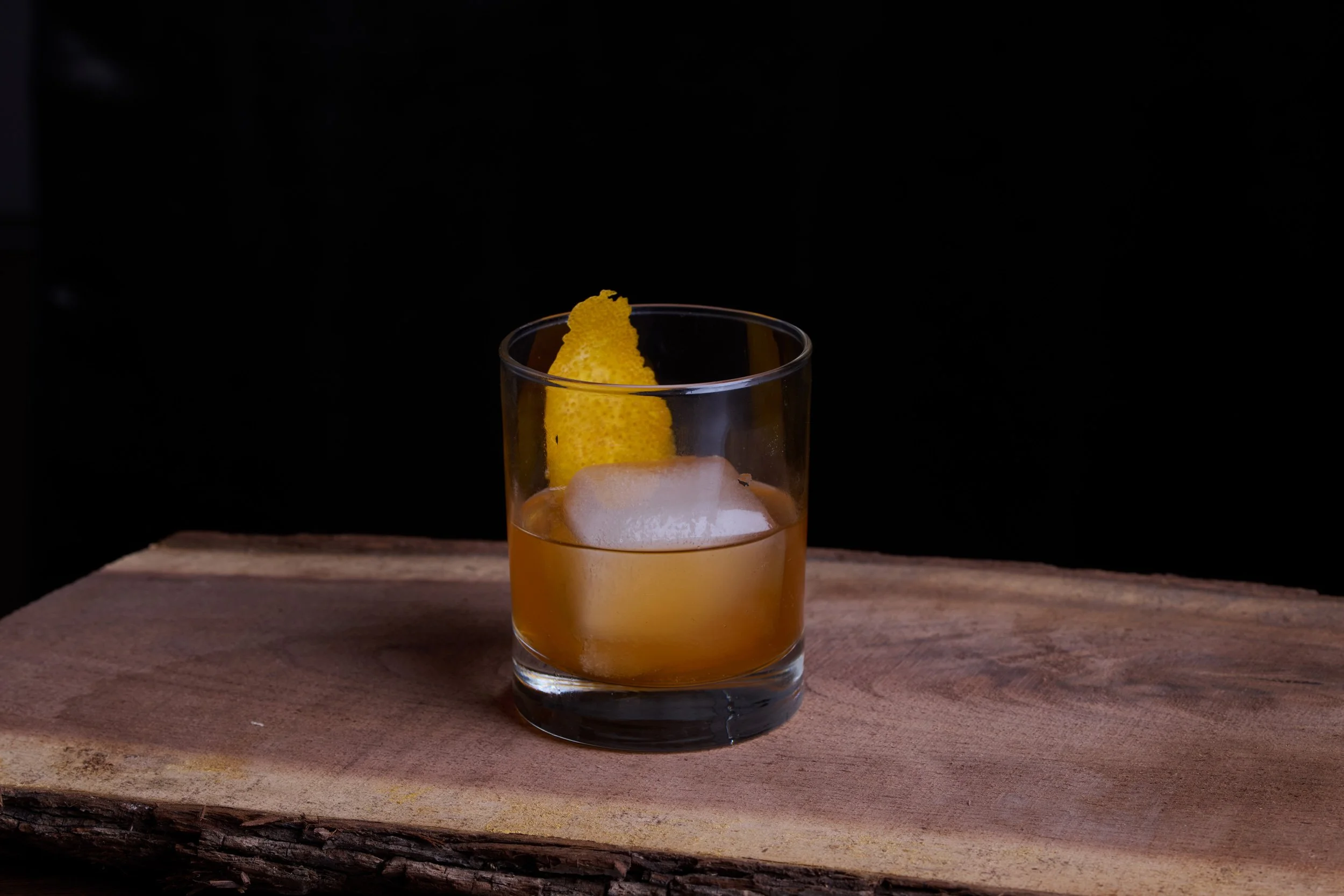 An unretouched shot of a glass of whiskey with a lemon peel garnish on a wooden surface against a black background.