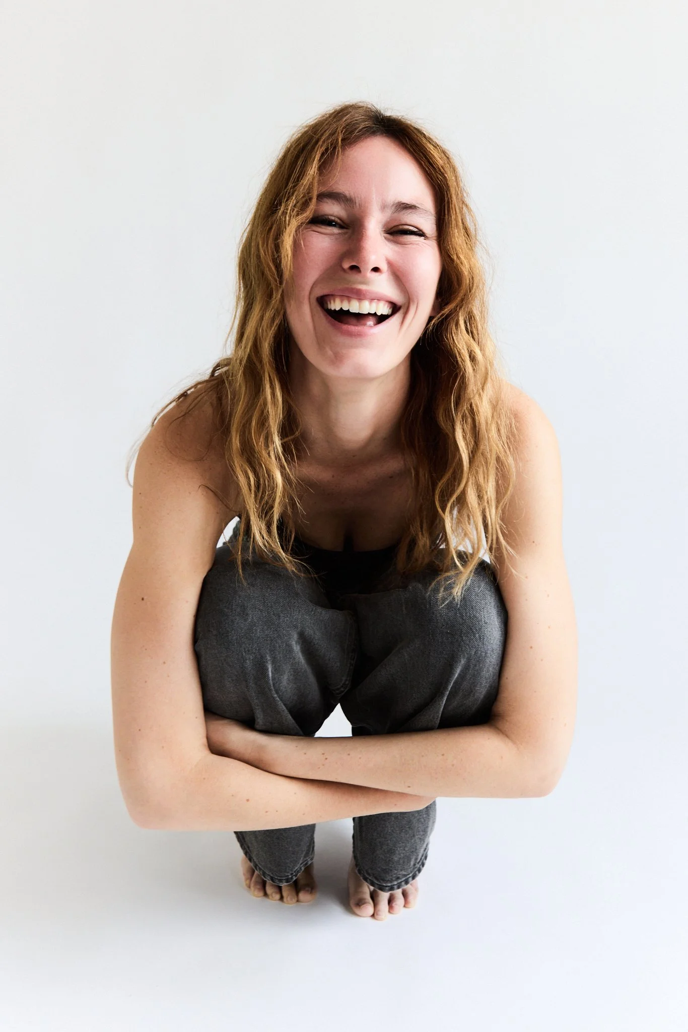 A woman with long wavy red hair, smiling and looking directly at the camera, is kneeling with her arms crossed in front of her, against a plain white background.