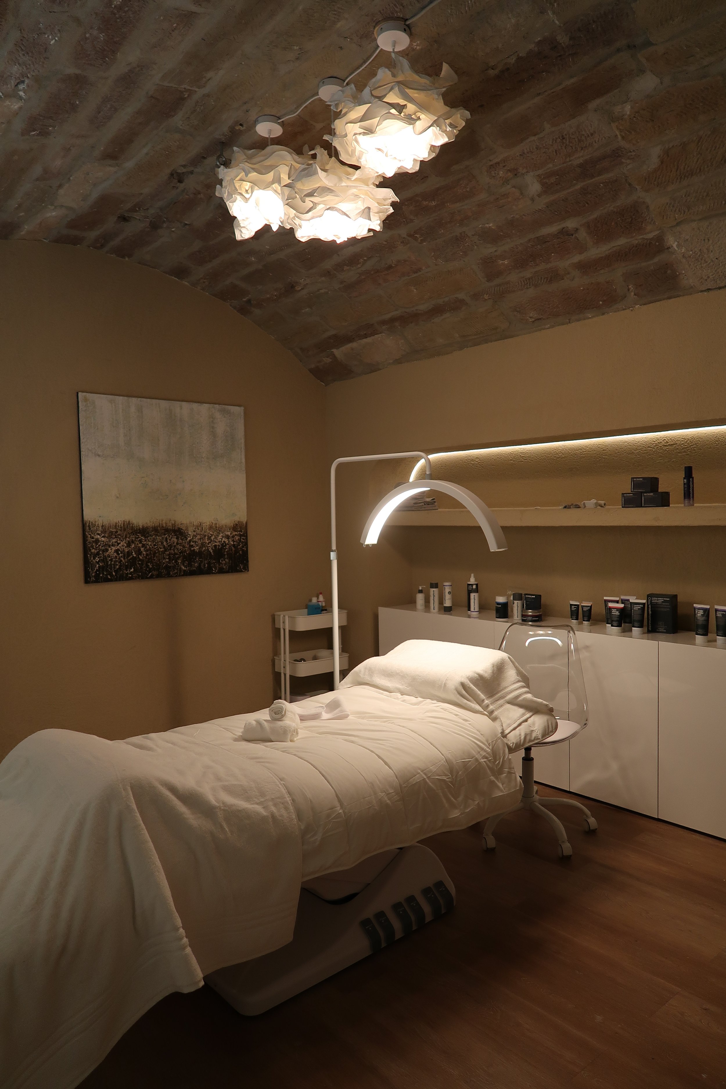 Ein Behandlungsraum mit einem Behandlungsbett, weißen Handtüchern, einer Stehlampe, einer Wandnische mit Pflegeprodukten, einem Wandbild und einem Deckenleuchter.