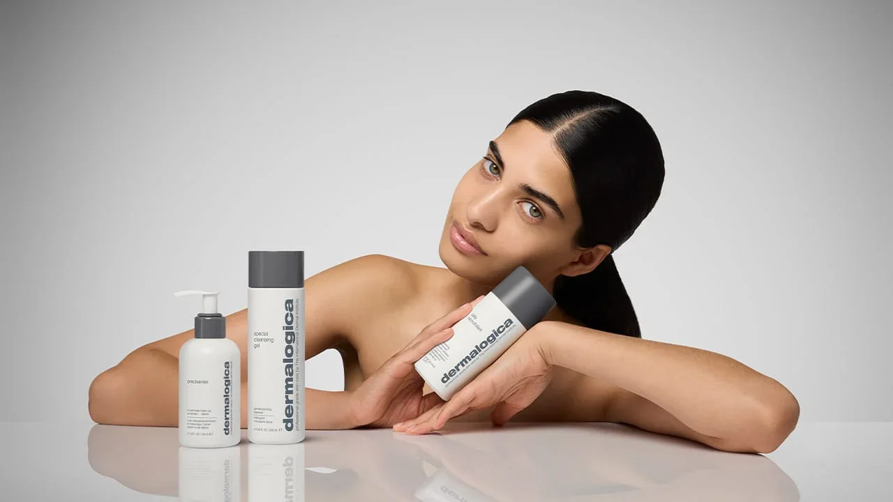 Eine junge Frau mit glatter, schwarzer Haarfrisur, die auf einem satinierten weißen Tisch liegt und drei Hautpflegeprodukte von Dermalogica präsentiert.
