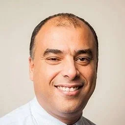 A man with short dark hair, smiling, wearing a light-colored dress shirt, against a plain background.