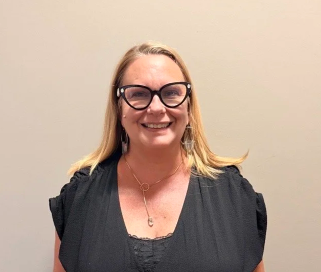 A woman with blonde hair wearing black glasses, earrings, a black top, and a necklace, smiling in front of a plain beige wall.