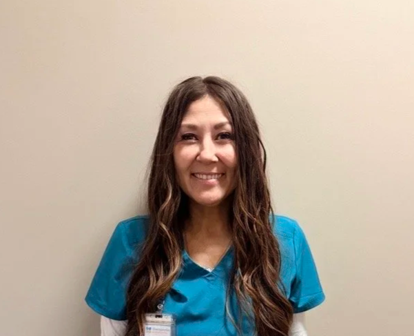 A woman with long, wavy brown hair wearing blue scrubs and a name badge, smiling against a plain beige wall.