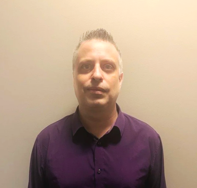 Man with short, spiked hair wearing a purple button-up shirt standing against a beige wall.
