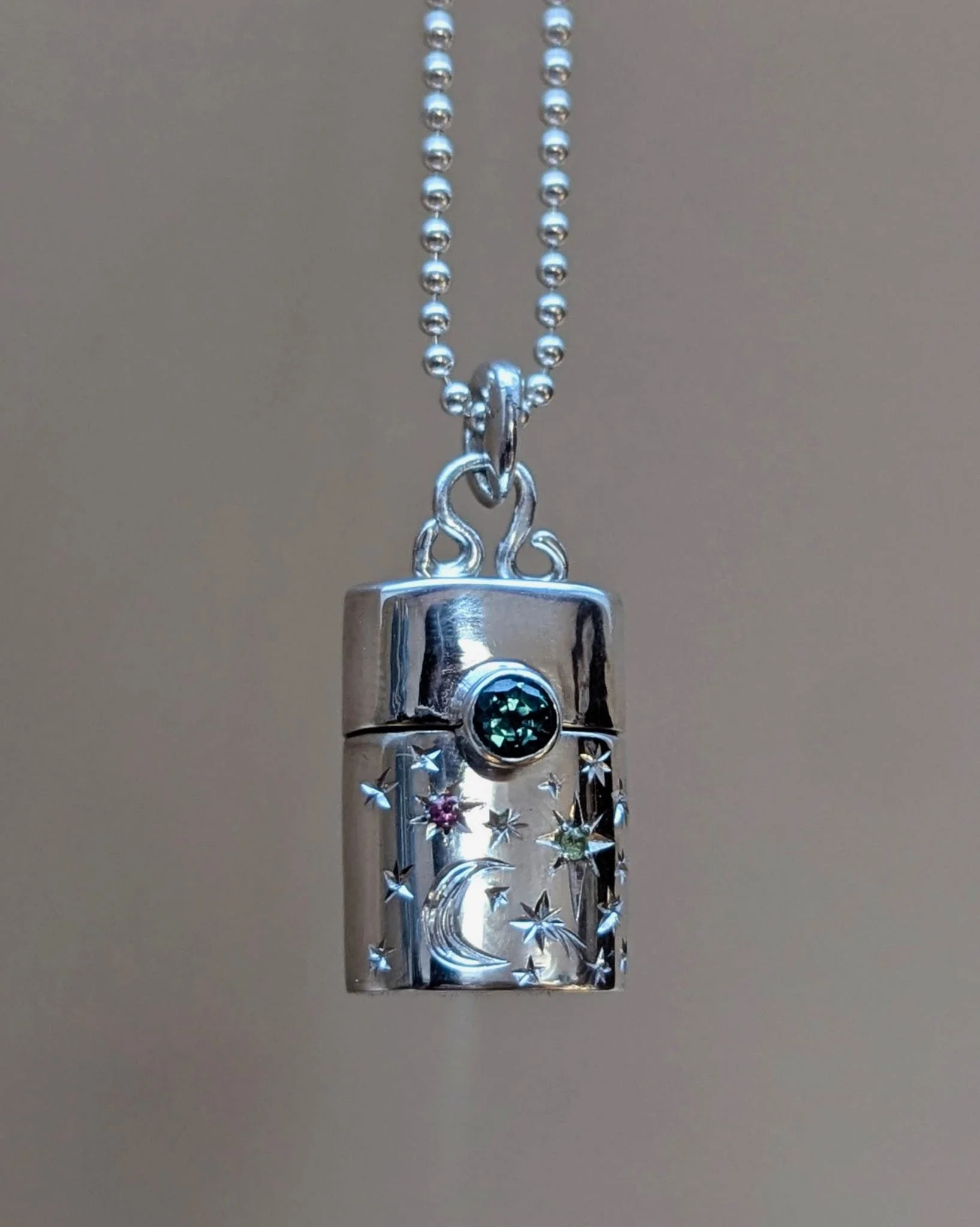 Close-up of a silver jewelry pendant hanging from a ball chain necklace, with decorative engravings and embedded gemstones, against a plain background.