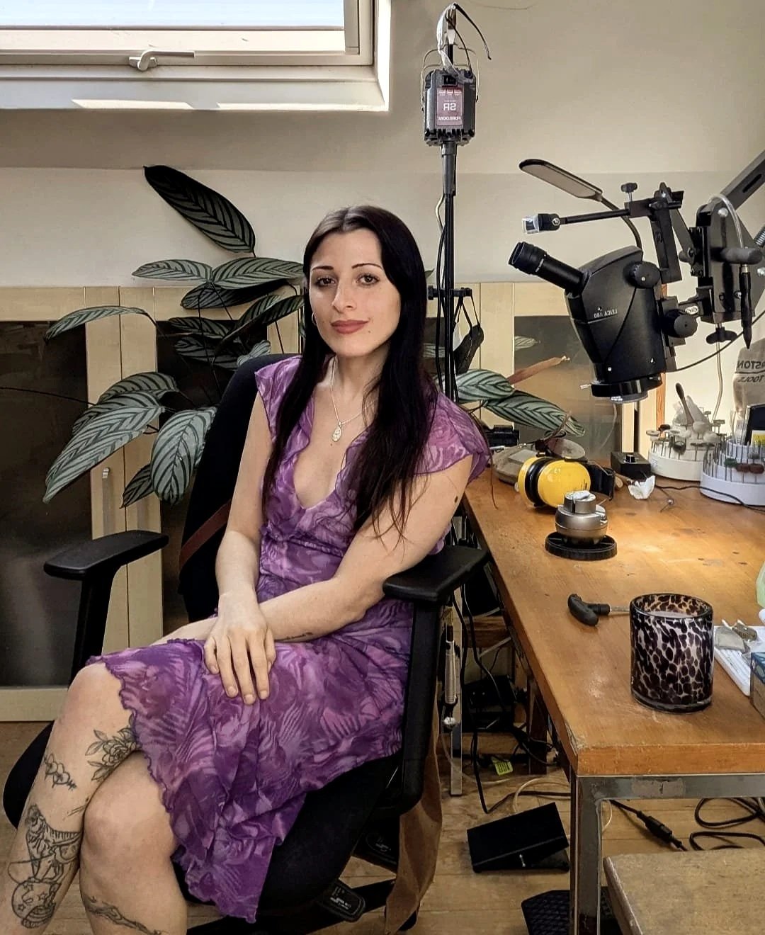 A woman with long dark hair sitting in a black chair in front of a wooden workbench. She is wearing a purple dress and has tattoos on her left arm and leg. The workspace contains a microscope, various tools, and equipment, with a large green plant in the background and a window overhead.